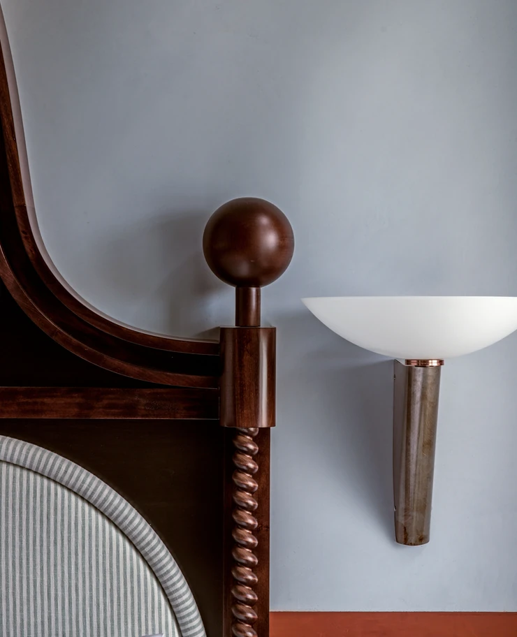 Close-up of a dark wooden bedpost with a rounded top and a twisted vertical design, next to a modern wall-mounted sconce with a white semi-circular shade on a blue-gray wall.