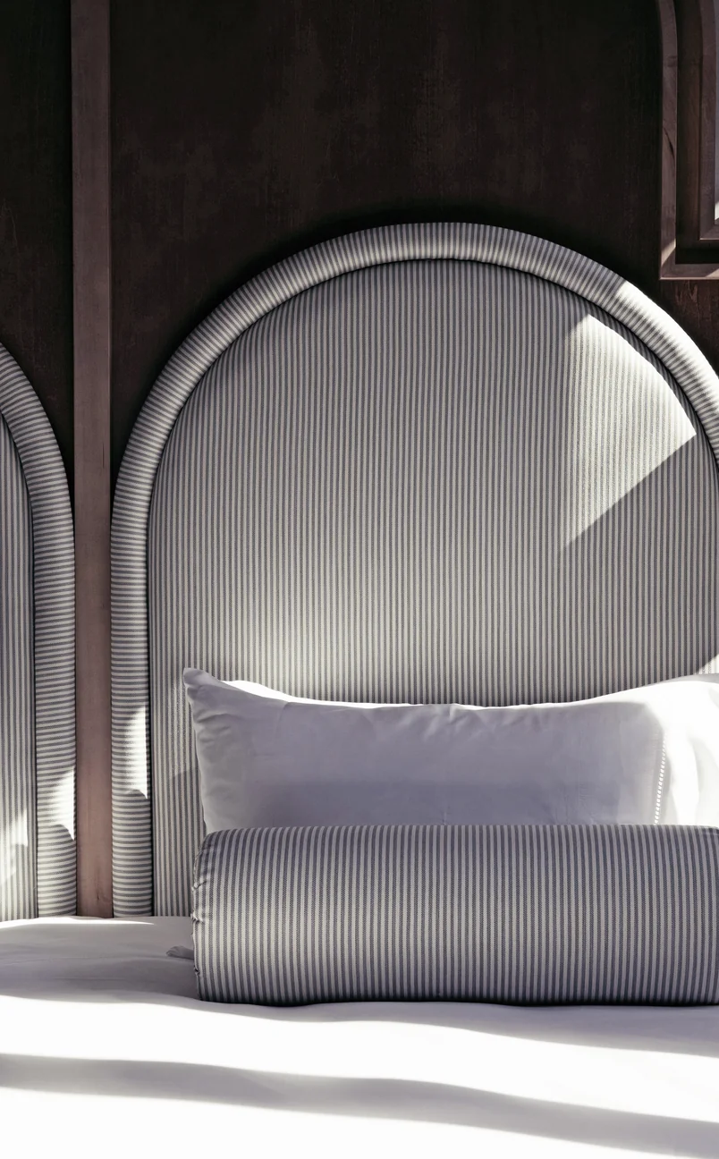 Close-up of a bed with a rounded, striped headboard, a white pillow, and a matching striped bolster pillow in soft natural light.