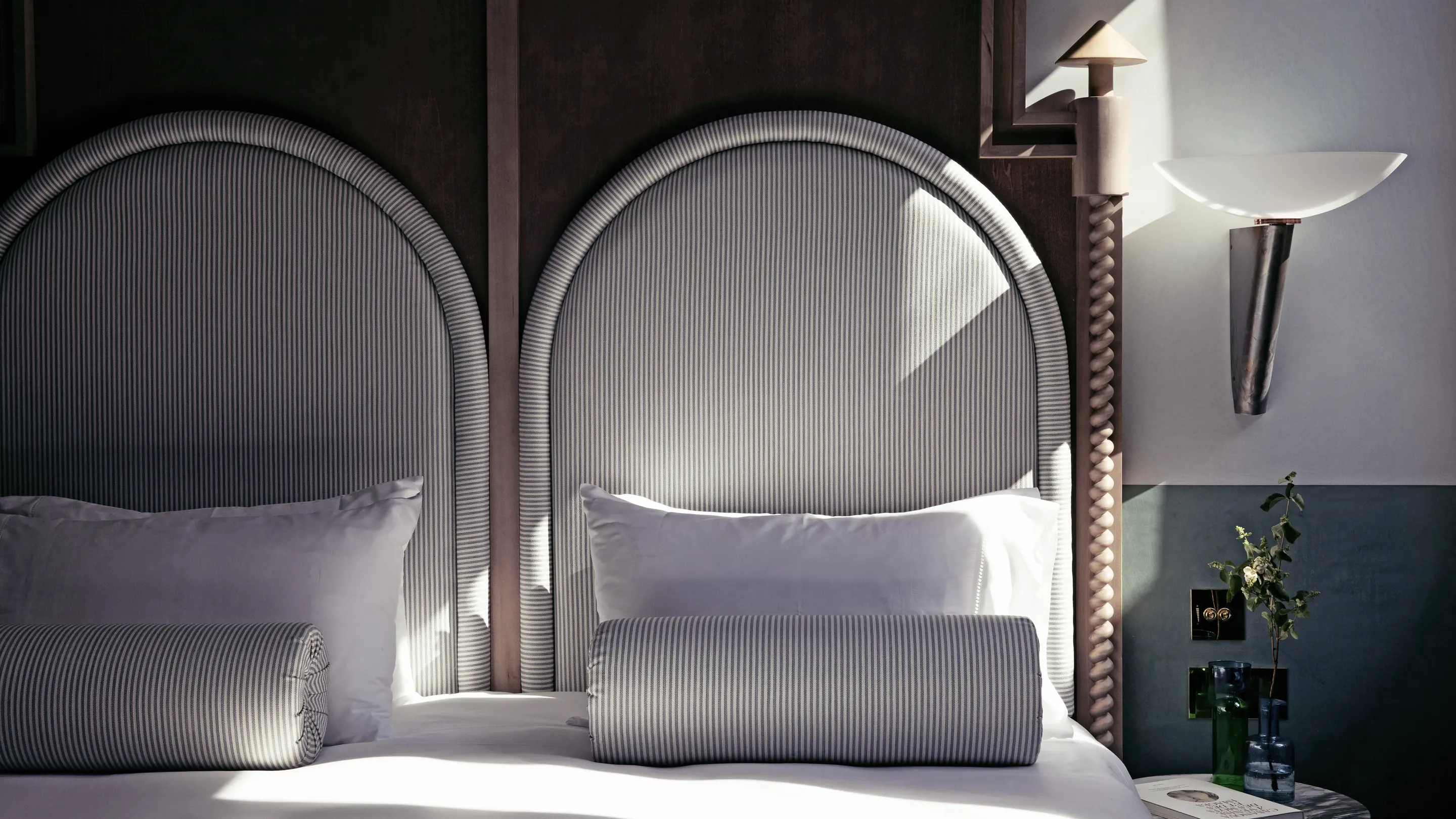 Bed with two gray striped upholstered headboards, white pillows, and cylindrical pillows, beside a small table with a plant and wall-mounted lamp.