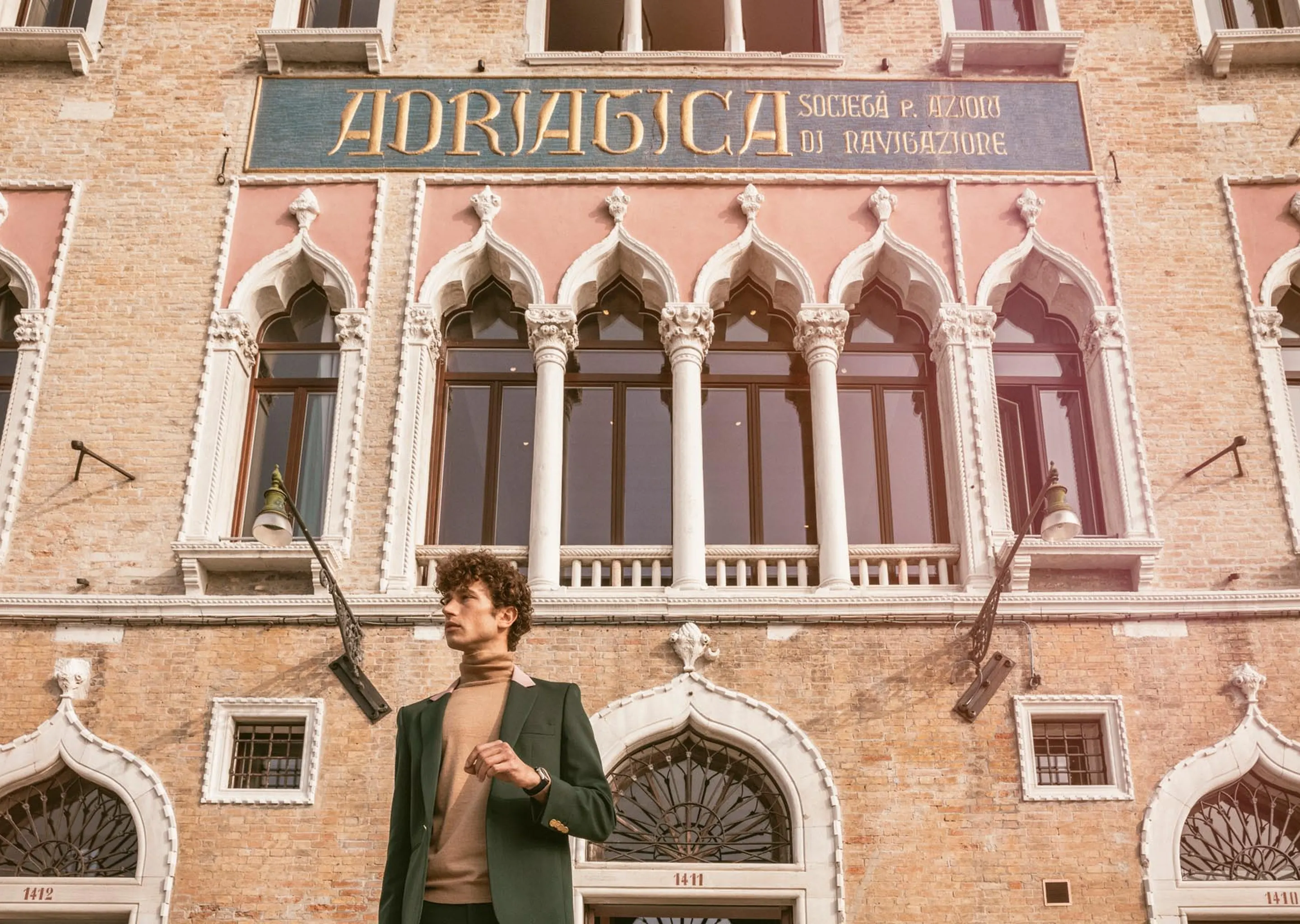 Man in a green suit and tan turtleneck standing in front of a historic brick building with arched windows and the sign 'ADRIATICA SOCIETÀ P. AZIORI DI NAVIGAZIONE'.