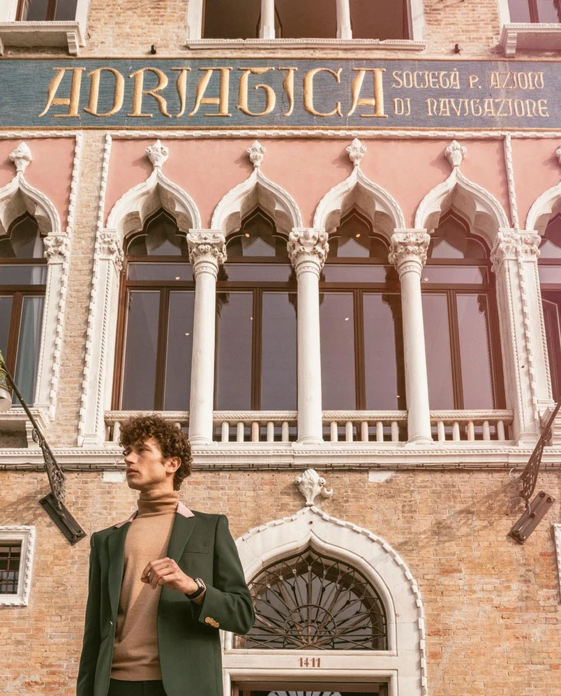 Man in green blazer and beige turtleneck standing in front of historic building with arched windows and Adriatica sign.