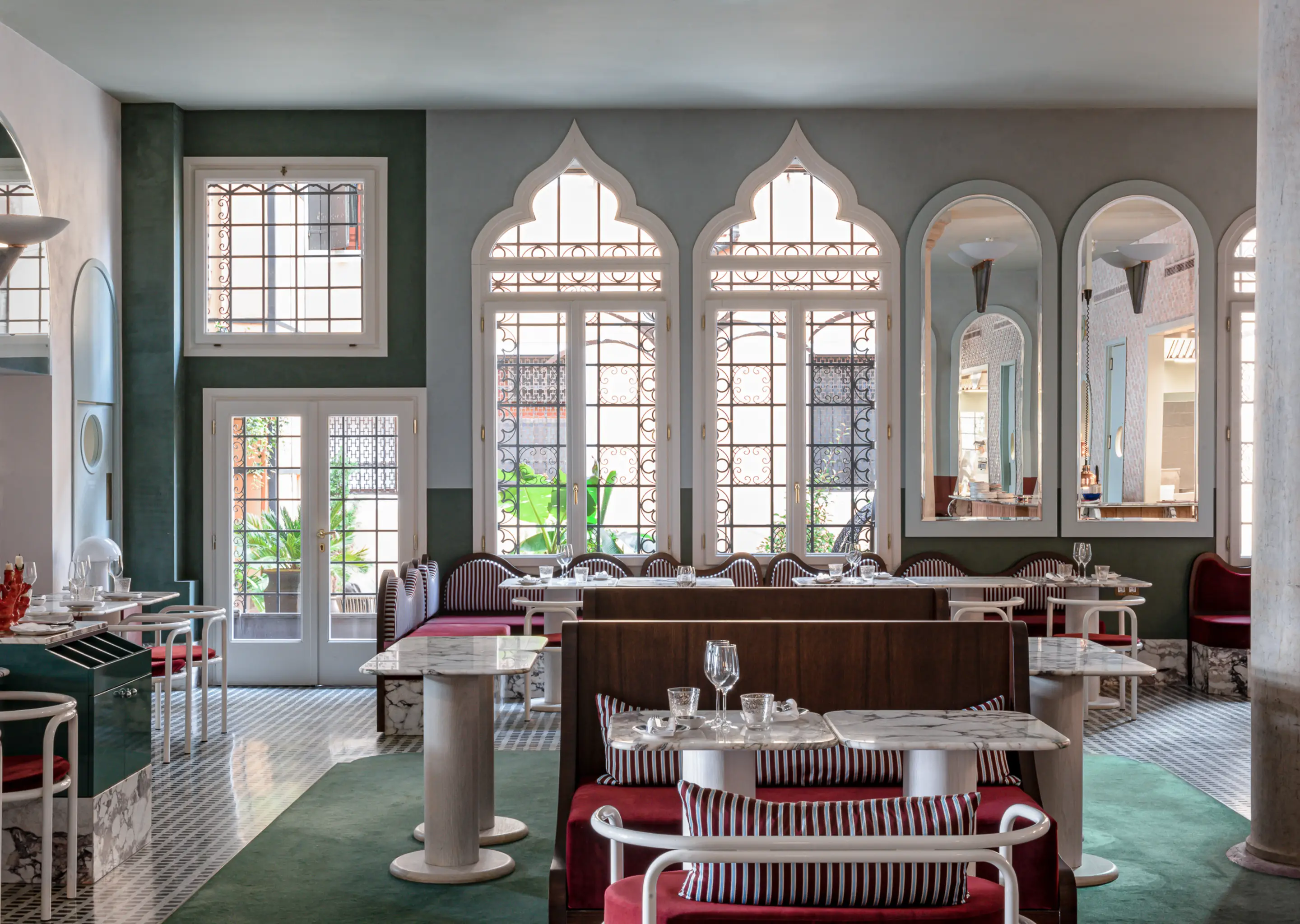 Elegant restaurant interior with marble tables, red cushioned seating, arched windows, and mirrors on the wall.