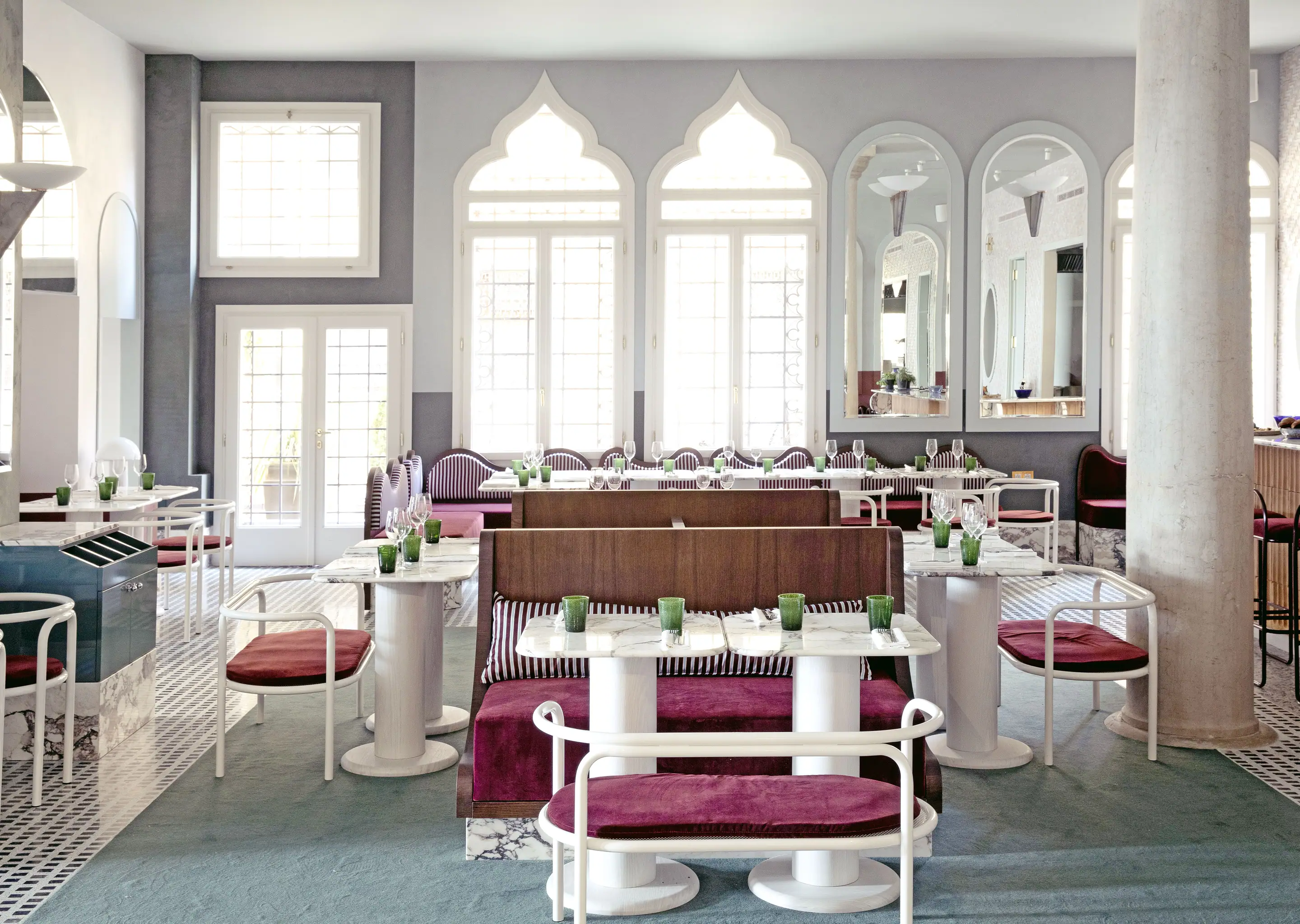 Bright hotel restaurant with arched windows, marble tables, purple cushioned seating, and green glassware.
