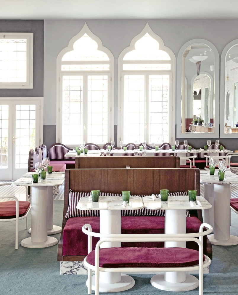 Elegant restaurant interior with purple velvet seating, marble tables, green glasses, large decorative windows, and wall mirrors.