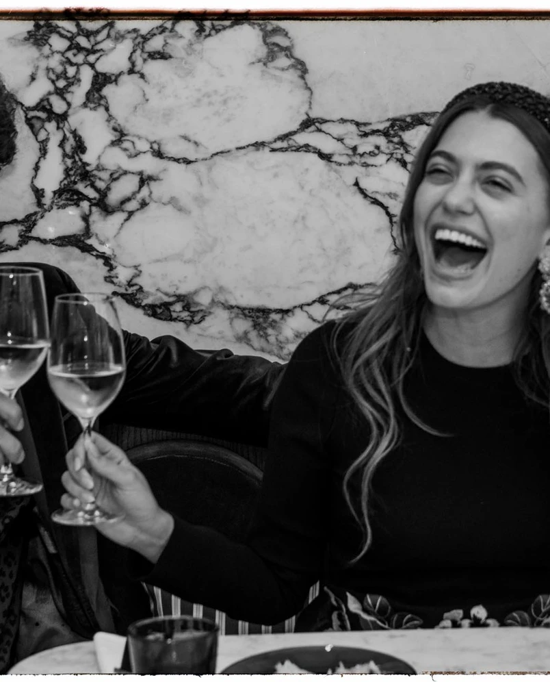 A woman laughing joyfully while holding a glass of white wine in a celebratory toast at a dining table.