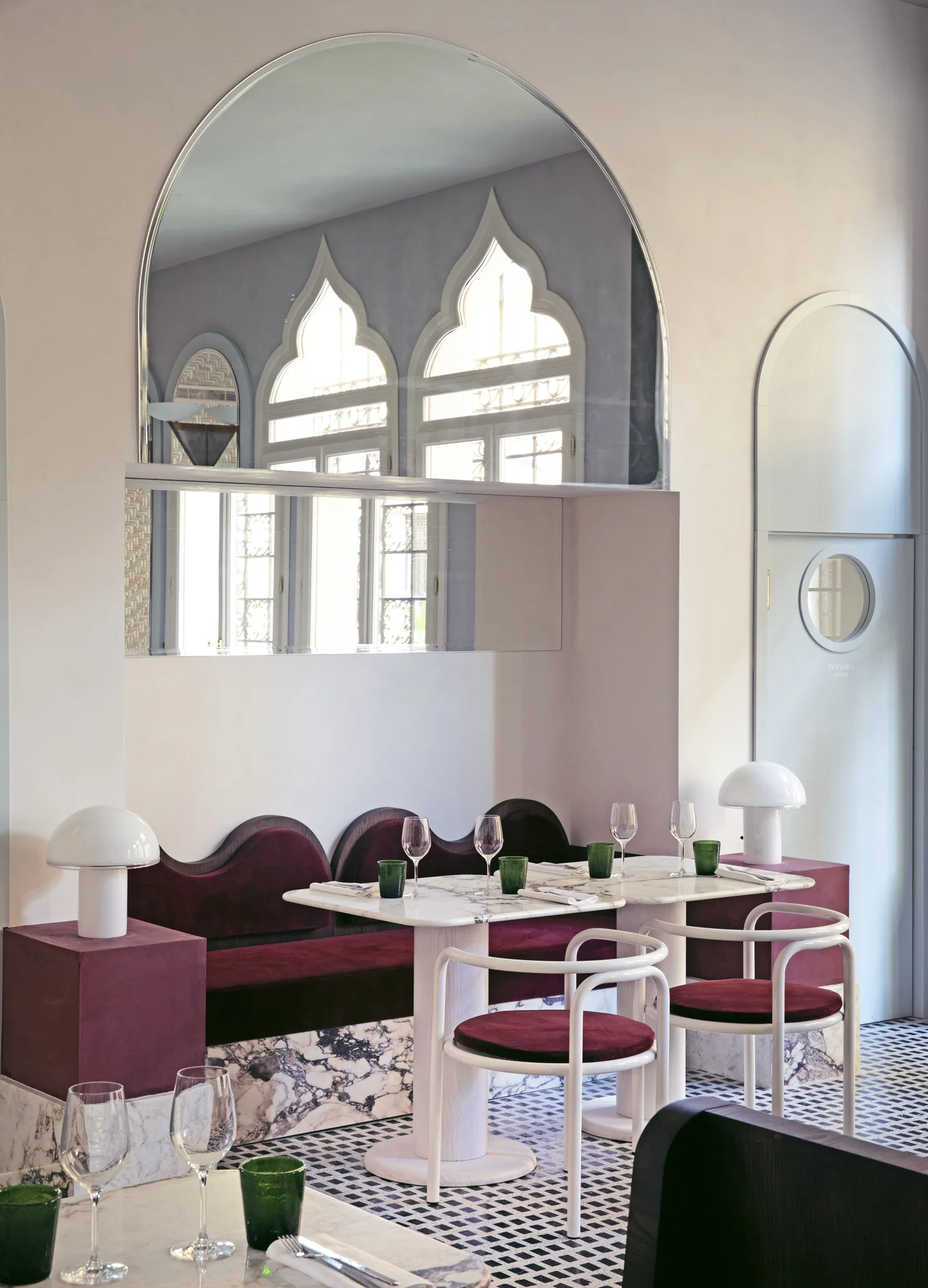 Modern dining area with marble tables, burgundy cushioned chairs and bench, decorative arched mirrors, and green glassware.