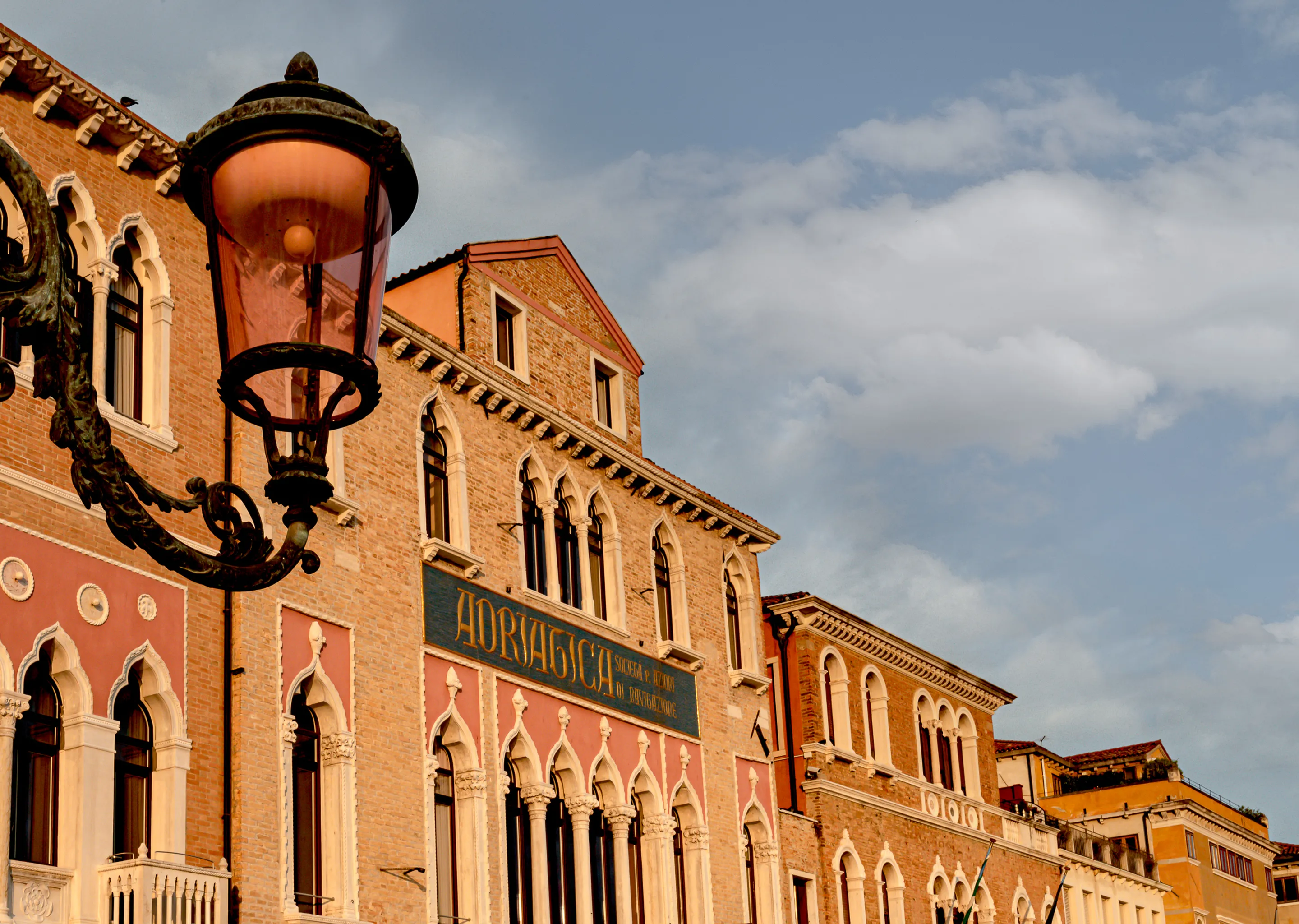 Historical brick buildings with arched windows under a partly cloudy sky and a large ornate street lamp in the foreground.