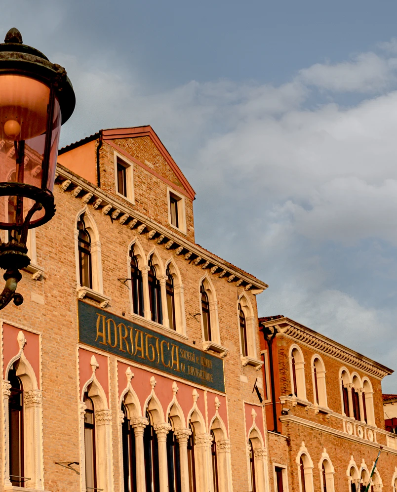Historic brick building with arched windows and a sign that reads Adriatica under a partly cloudy sky, with an old street lamp in the foreground.