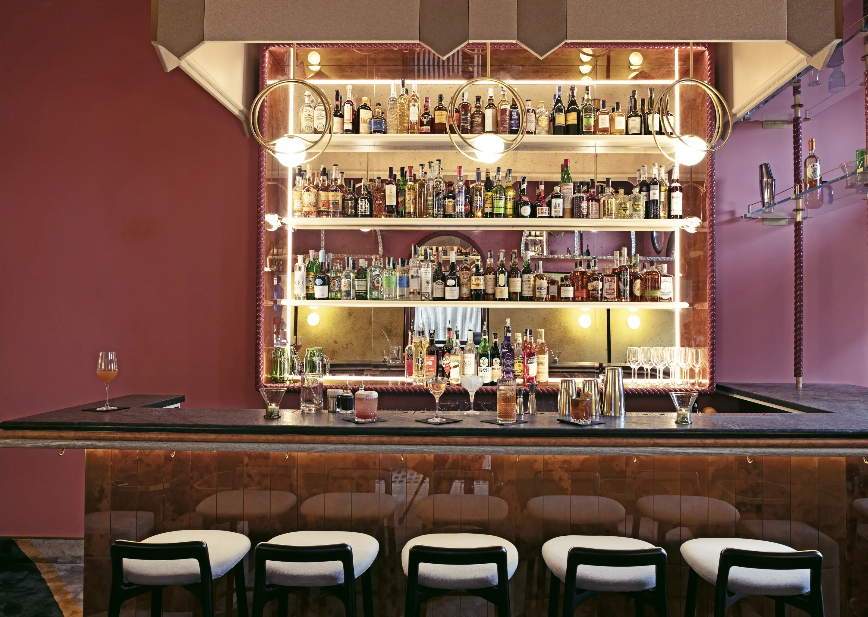 Elegant cocktail bar with illuminated shelves stocked with various liquor bottles, modern round pendant lights, and six cushioned stools along the marble counter.