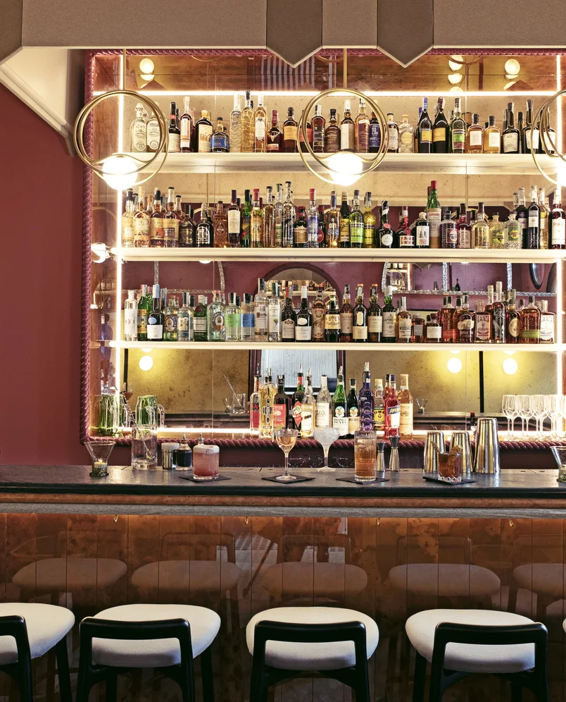 Modern hotel cocktail bar with illuminated shelves of various liquor bottles, hanging globe lights, and four bar stools with white seats.