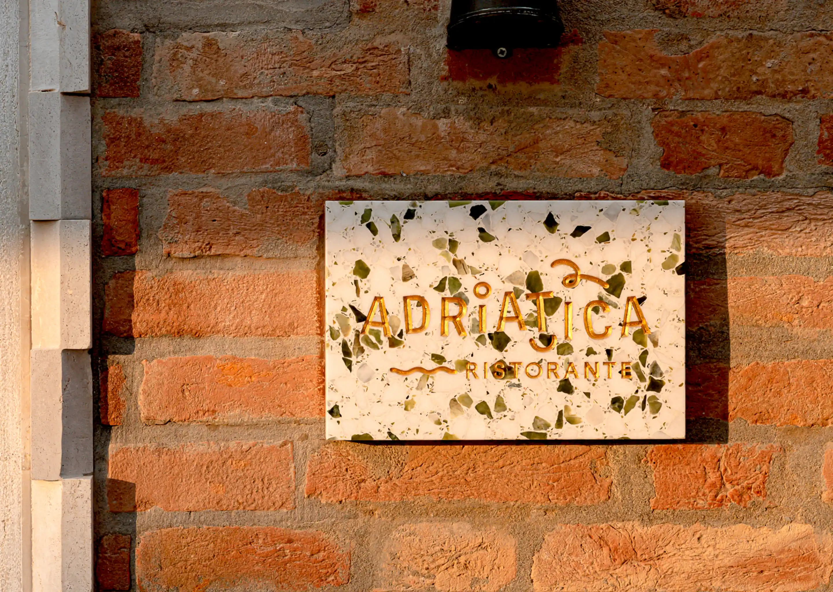 Wall-mounted terrazzo sign with the text 'Adriatica Ristorante' in gold letters on a brick wall.