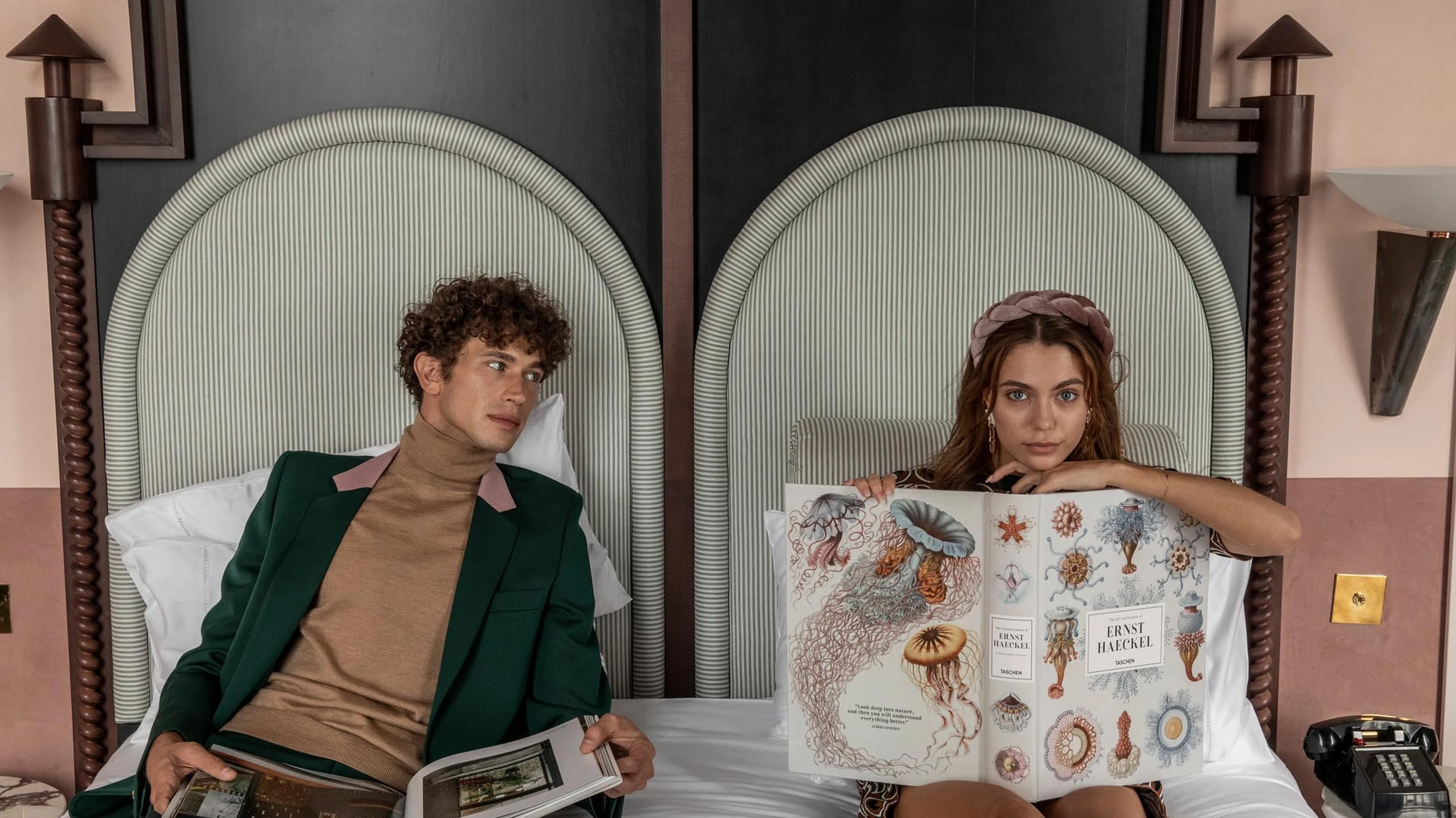 Hombre con traje verde y cuello alto marrón y mujer con una diadema rosa sentados en las camas leyendo libros ilustrados.
