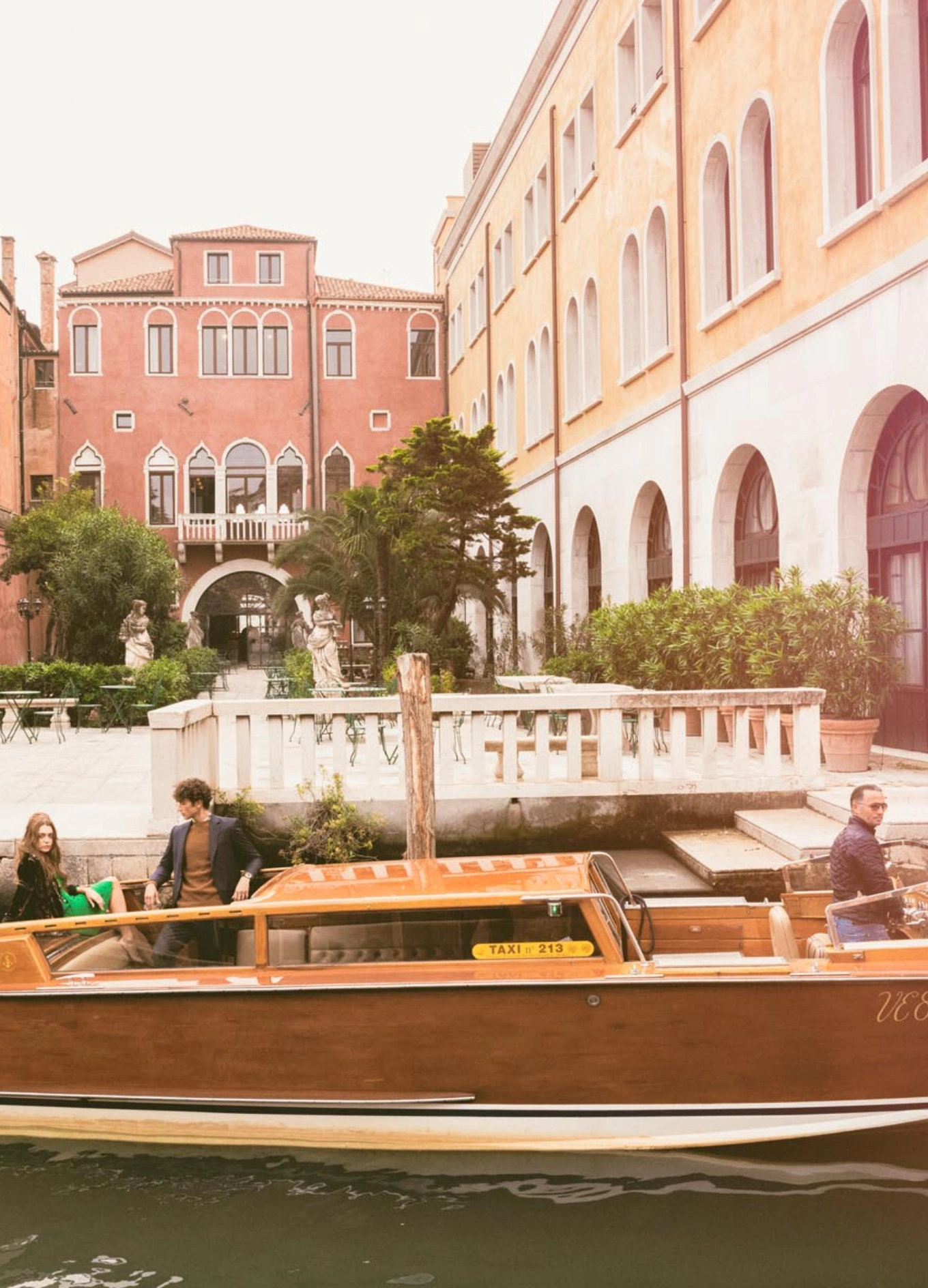 Wooden water taxi with three people on a canal next to buildings with arched windows and greenery.