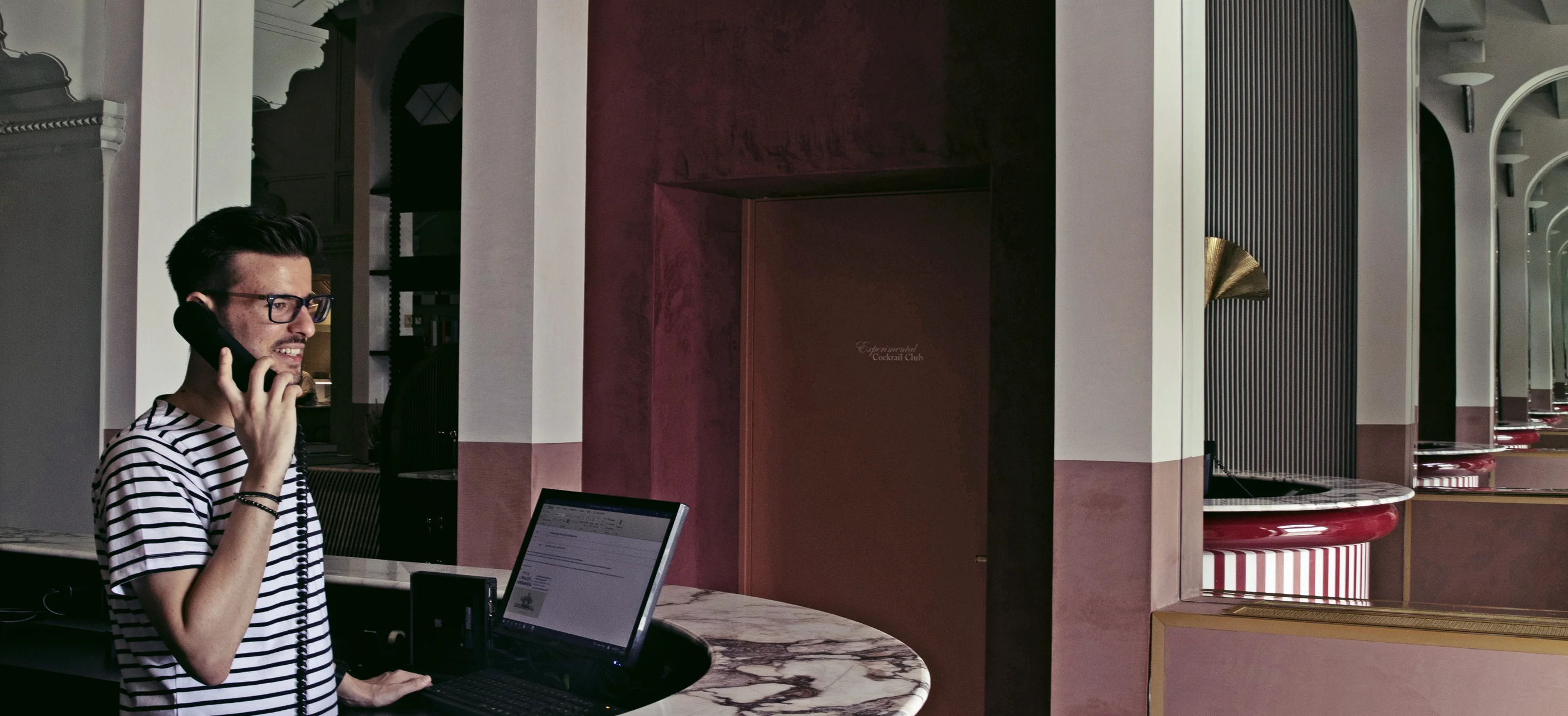 Man in glasses and striped shirt speaking on a corded phone behind a marble counter with a computer in a stylish interior.