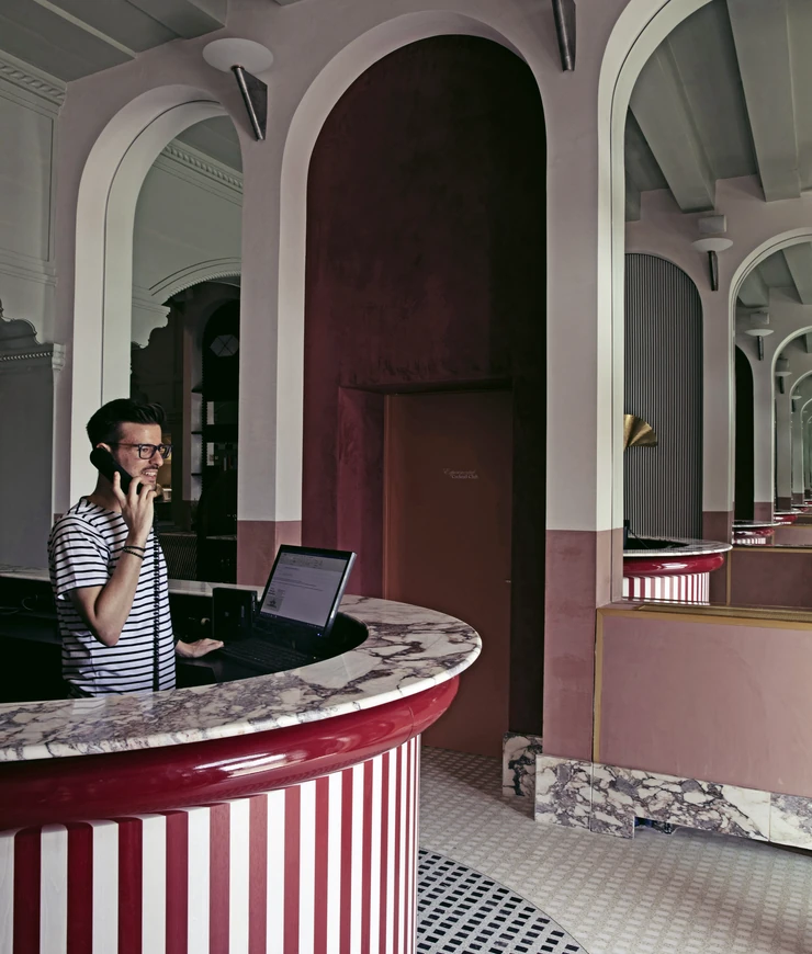 Man in striped shirt talking on a black phone at a round reception desk with red and white stripes and marble countertop inside a hotel with arched mirrors.