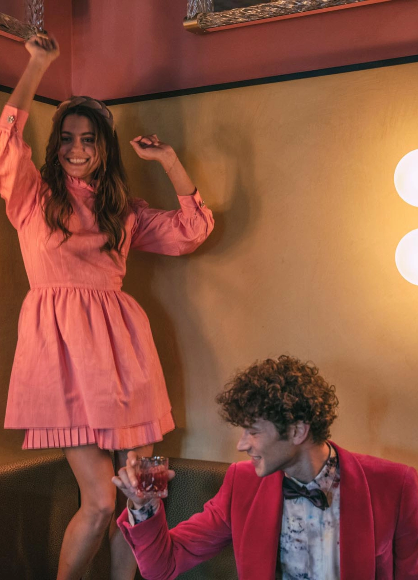 A woman in a pink dress dancing joyfully while a man in a pink jacket and bow tie holds a drink and watches her, both in a warmly lit room.