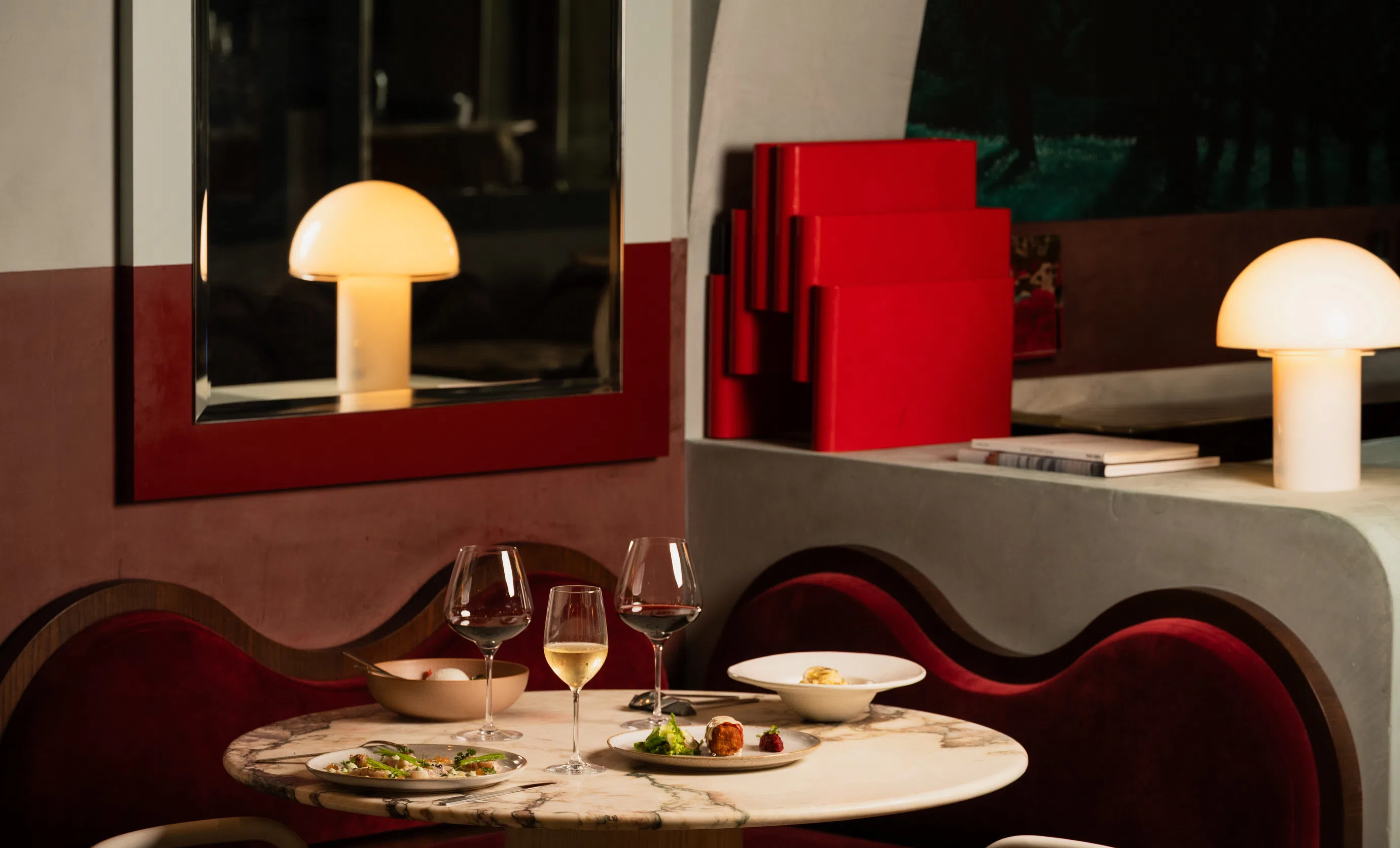 Marble table in a restaurant with plated gourmet dishes, wine glasses, and warm mushroom-shaped table lamps.