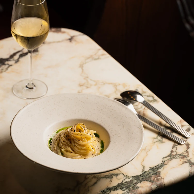 Elegant plate of creamy pasta garnished with herbs and black pepper on a marble table next to a glass of white wine and silverware.