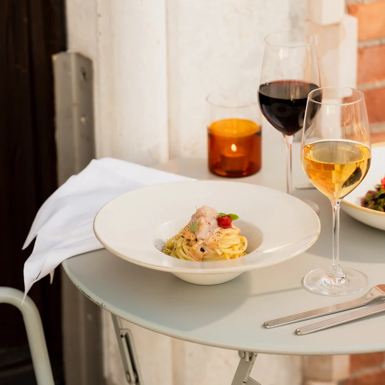 White plate with pasta topped with sauce and herbs on a round gray table with a glass of white wine, a glass of red wine, and an orange candle in the background.