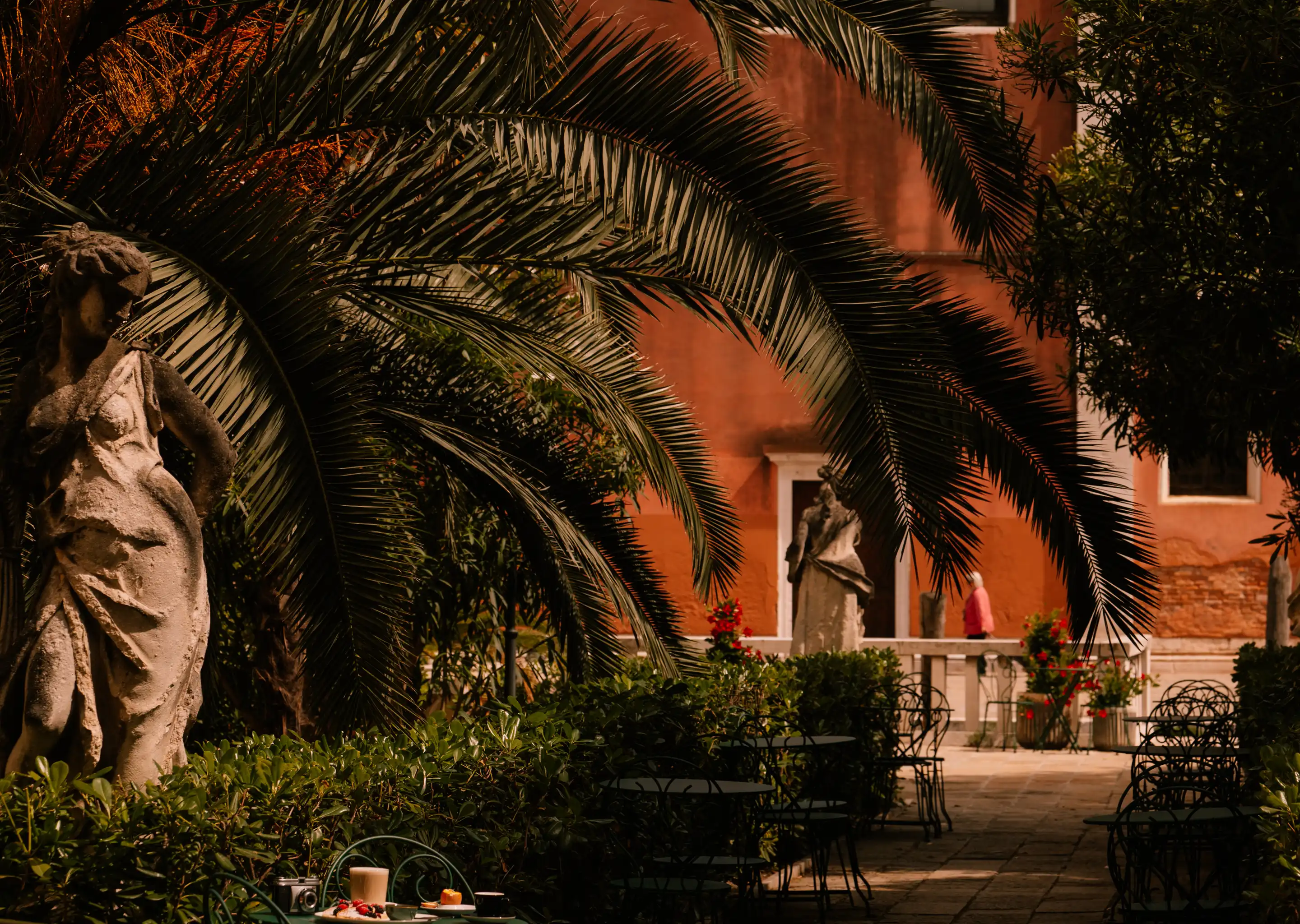 Shaded garden courtyard with palm leaves, stone statues, tables, chairs, and a person walking near an orange building.