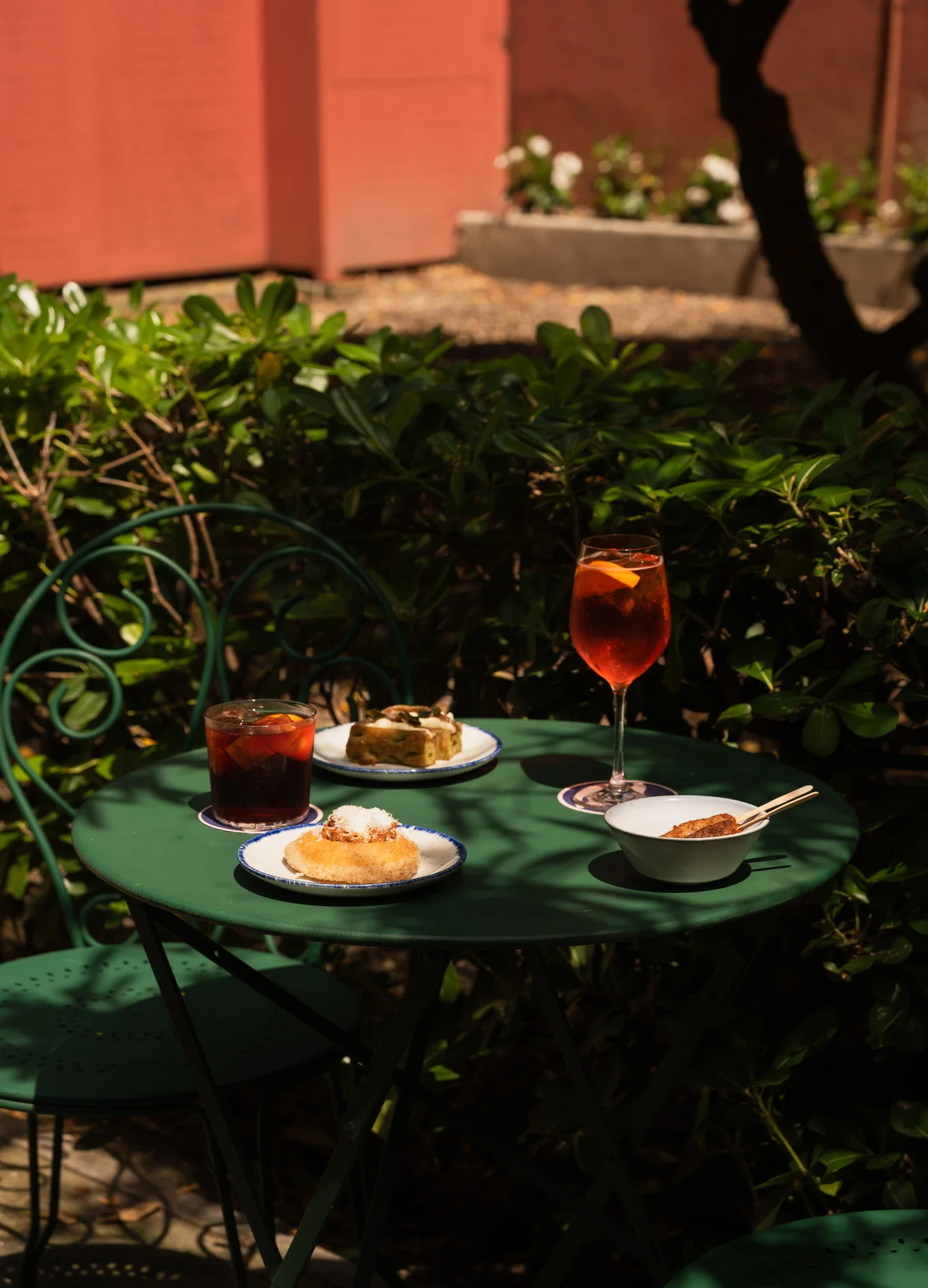 Green outdoor table with two drinks and assorted appetizers in a garden setting with green foliage.