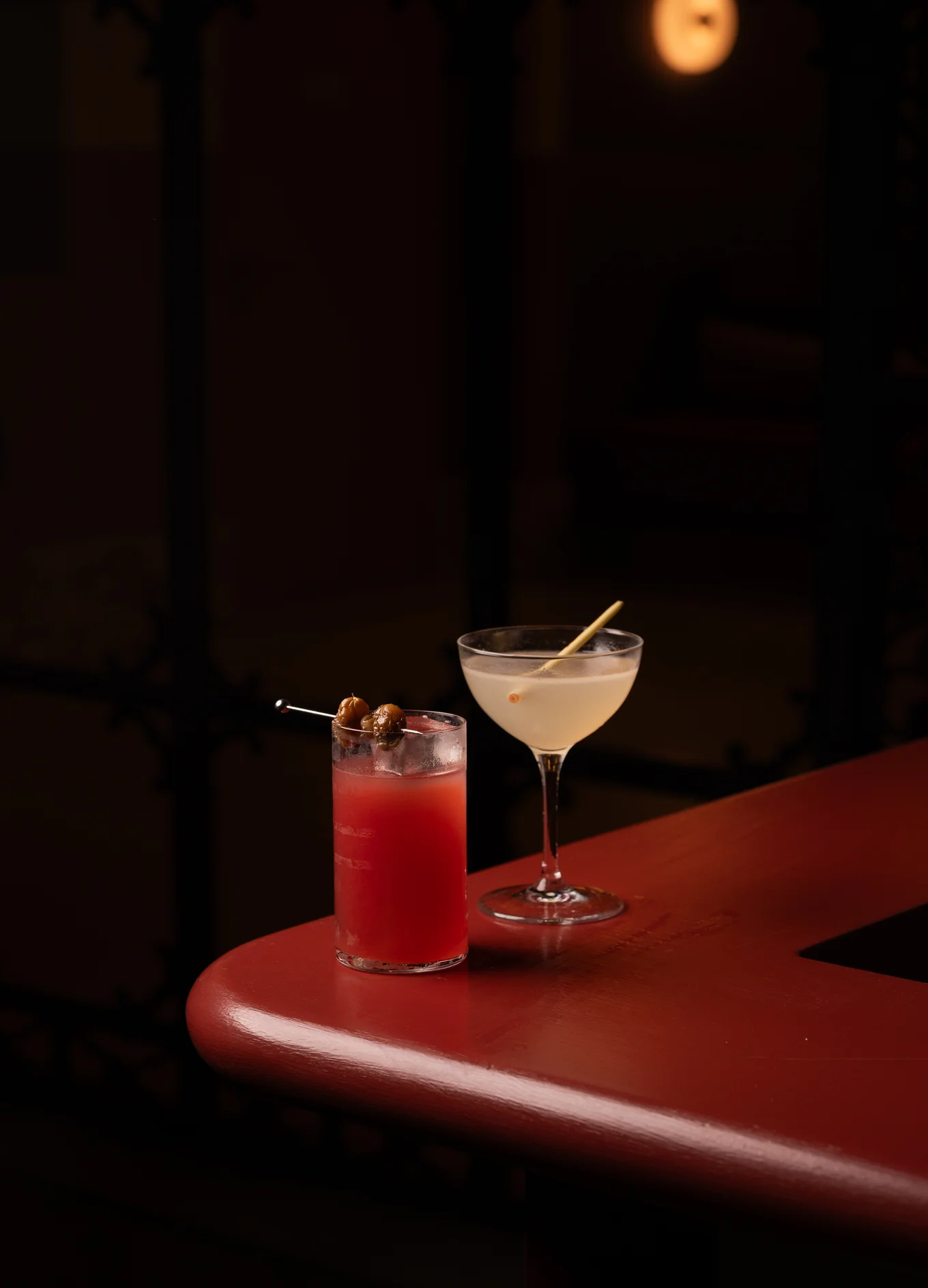Two cocktails on a red bar counter, one red drink with ice and olives, and one pale drink with a lemongrass garnish.