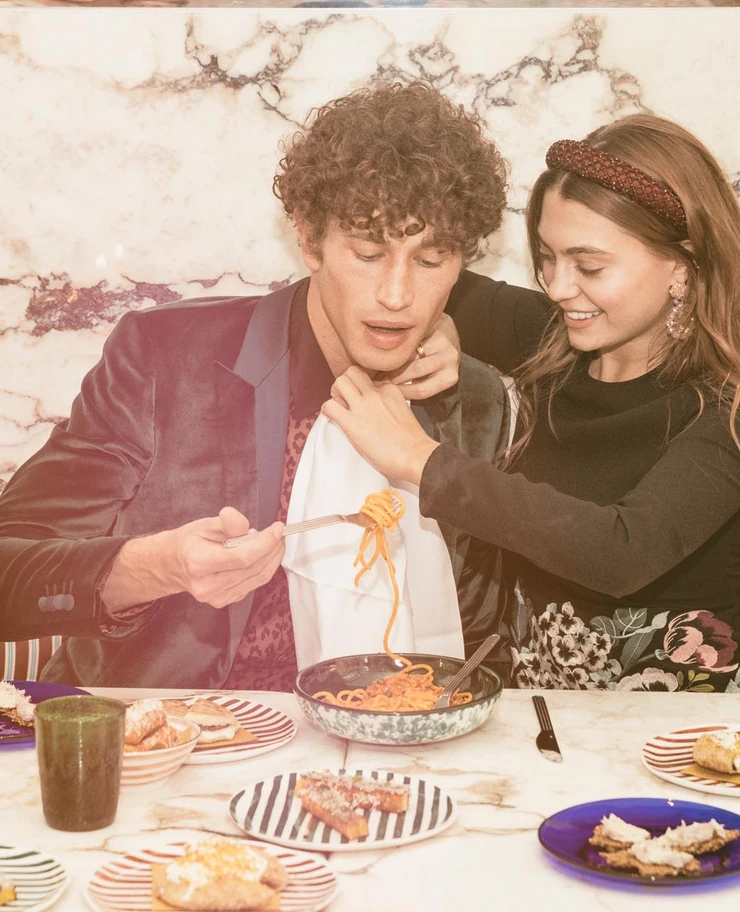 A woman helping a man put on a bib while he is holding a fork with spaghetti over a bowl at a table with plates of food.