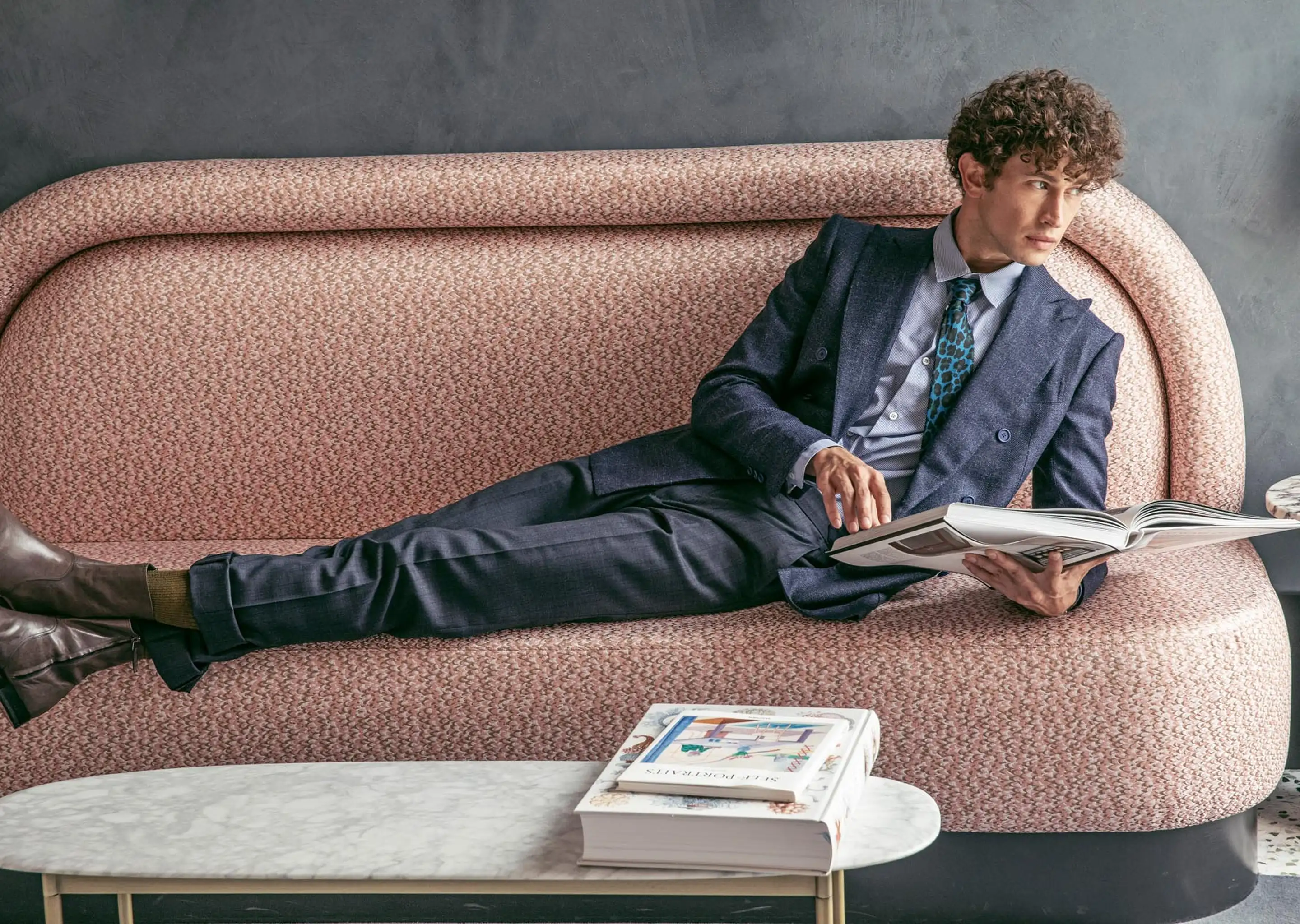 Man in a dark suit with a patterned tie lounging on a pink textured sofa while reading a large book.
