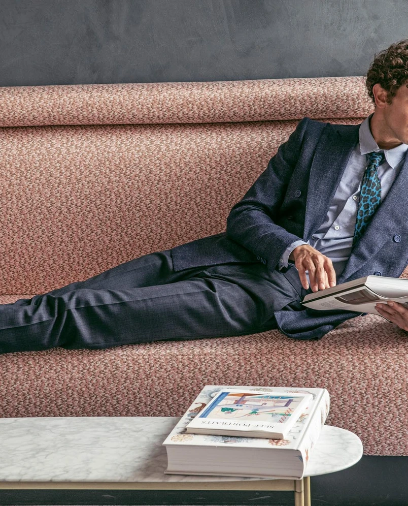 Man in a dark blue suit with a patterned blue tie reclining on a textured pink couch, reading a book, with a marble coffee table in front holding a large book.