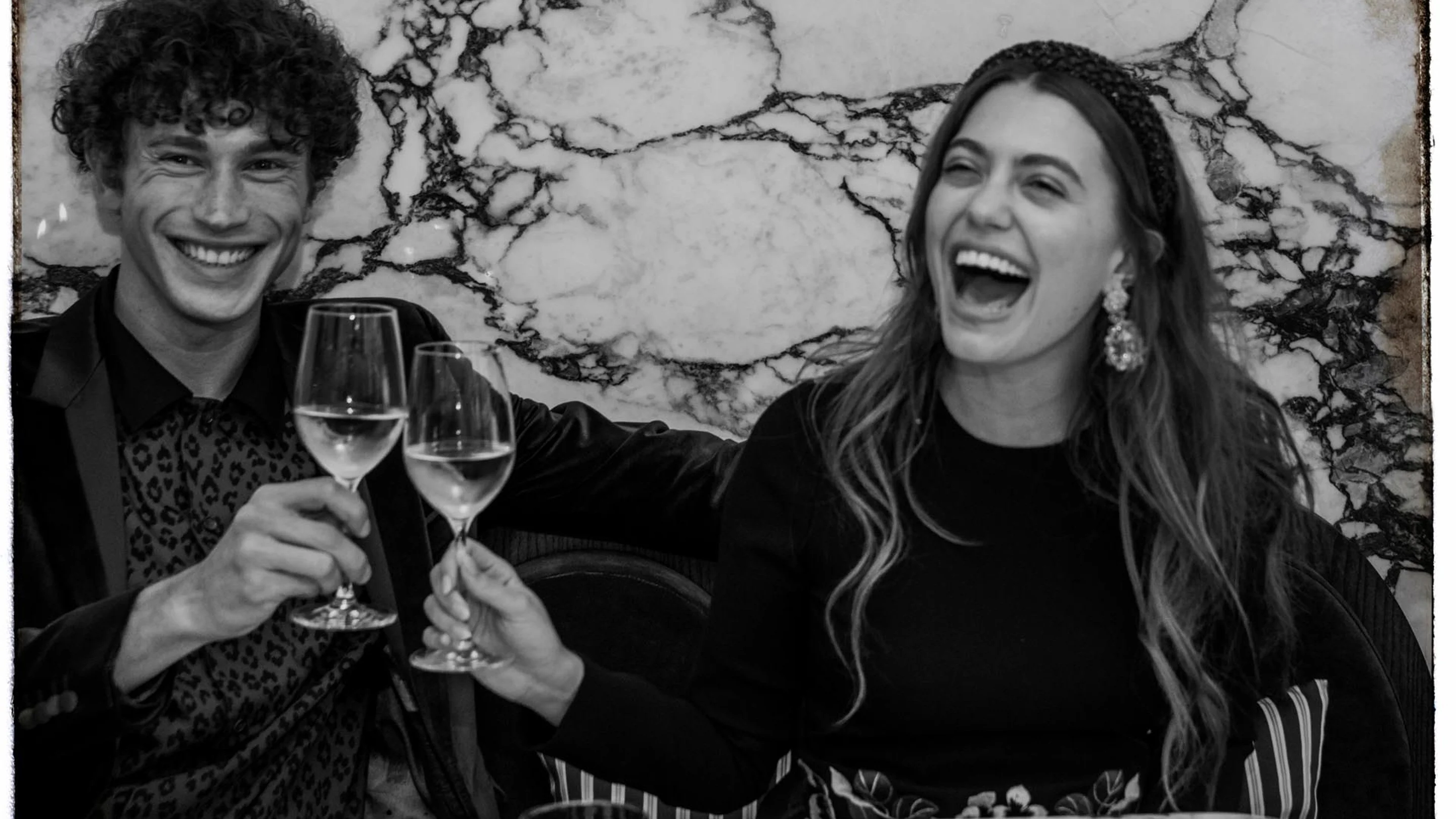 Smiling man and laughing woman clinking wine glasses against a marble wall background.