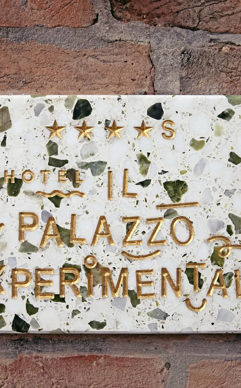 Hotel Il Palazzo Experimental sign with four gold stars on a white terrazzo background with green and gray stones, mounted on a brick wall.