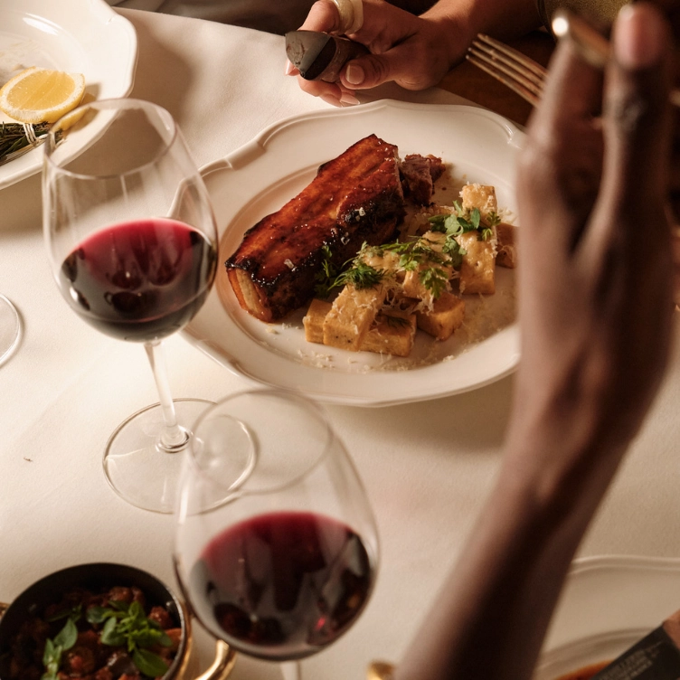 Plate with a piece of glazed meat and garnished potatoes, accompanied by two glasses of red wine on a white tablecloth.