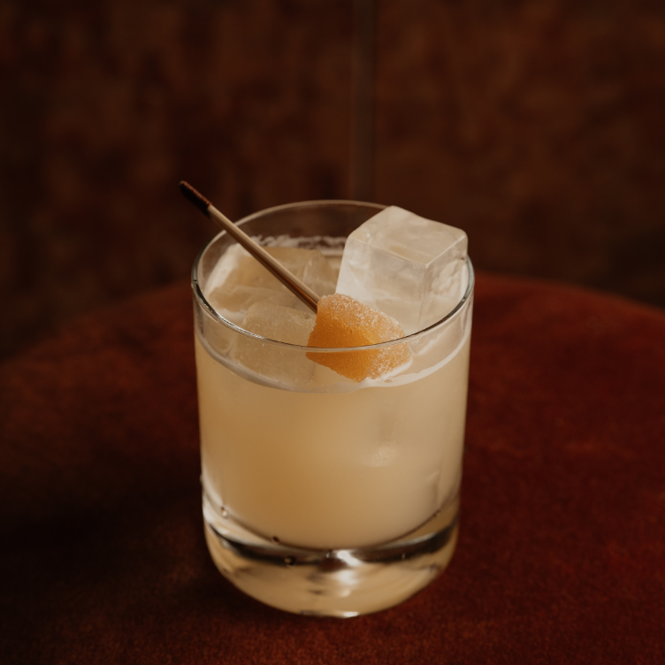 Cocktail in a clear glass with ice cubes and a small orange peel on a cocktail stick, set on a brown surface.