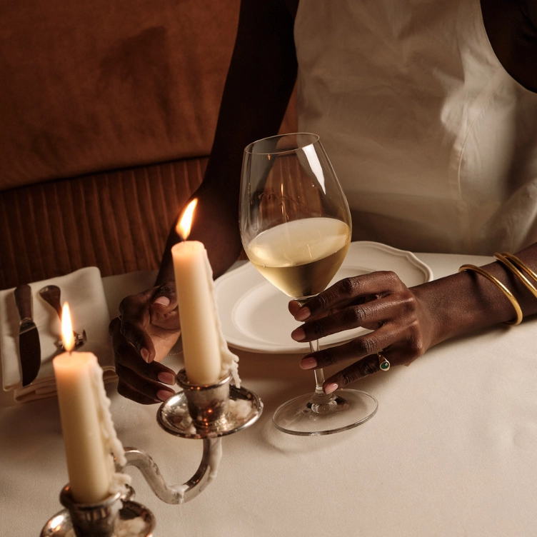Person with dark skin holding a glass of white wine next to two burning candles on a white tablecloth.