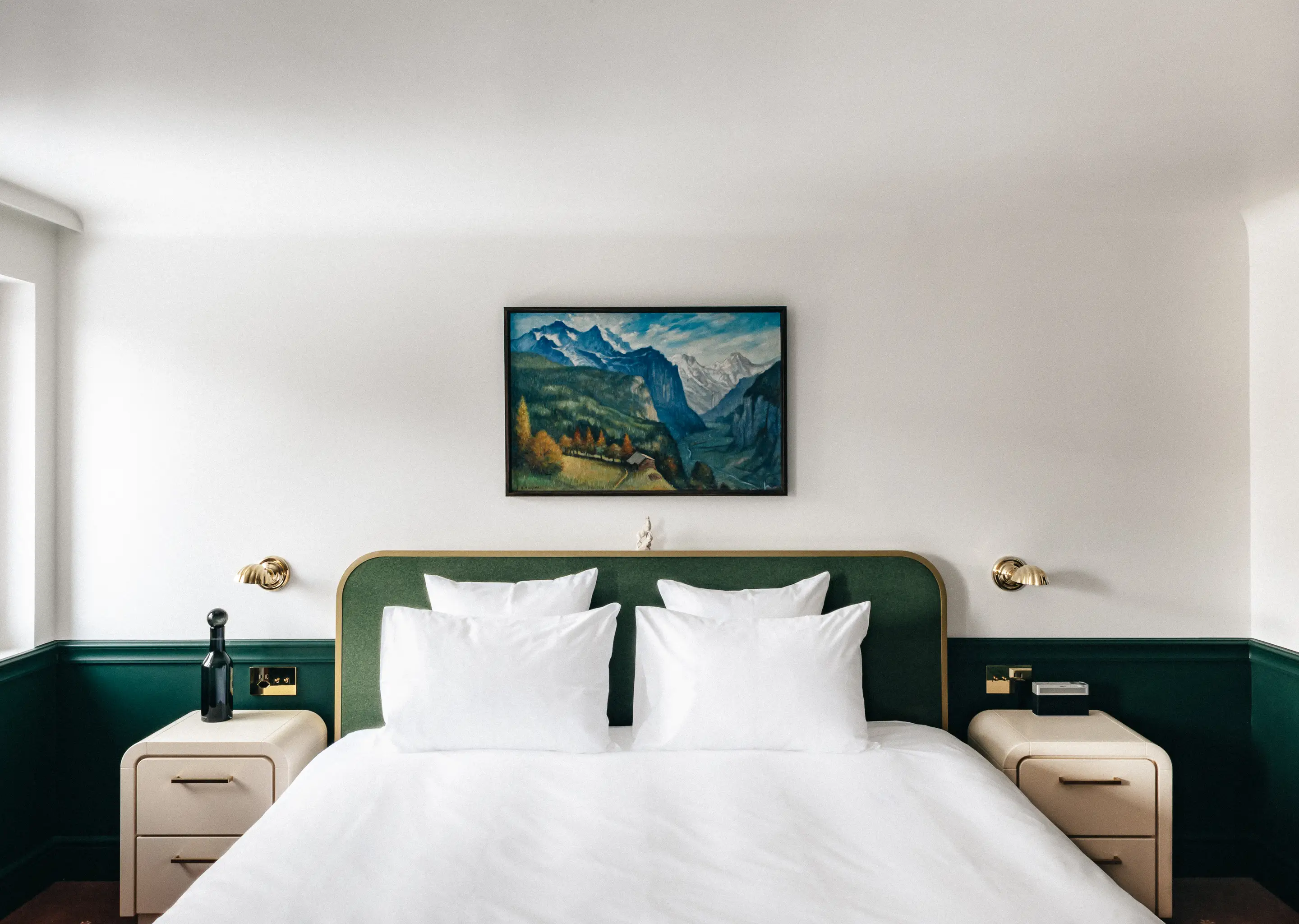 Modern hotel room with a white bed, green headboard, two bedside tables, and a mountain landscape painting above the bed.