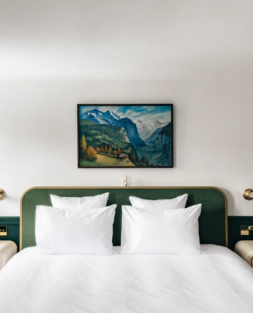 Modern hotel room with a green upholstered headboard, four white pillows, and a landscape painting of mountains above the bed.