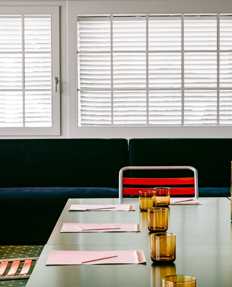 Modern dining table with amber glasses, pink napkins, and red-striped chairs in front of white window blinds and a dark couch.