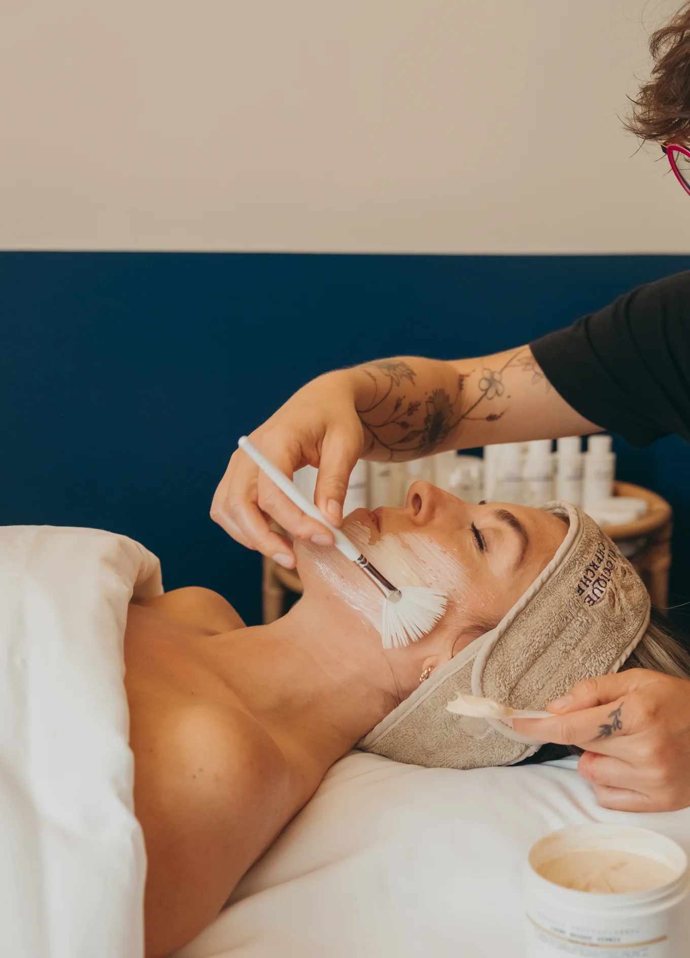 Person receiving a facial treatment with a cream being applied to their cheek using a fan brush by a tattooed esthetician.