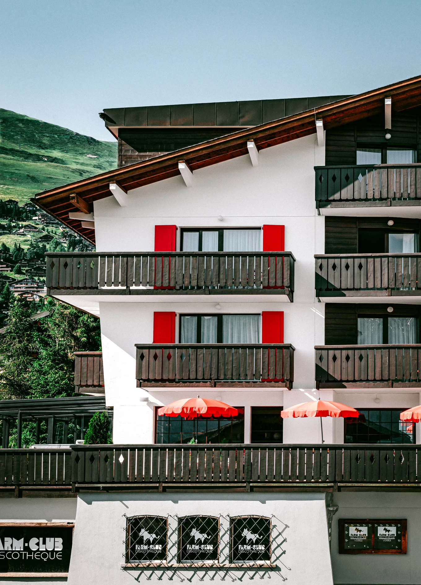 Exterior of a chalet-style building with wooden balconies, red window shutters, and red patio umbrellas.
