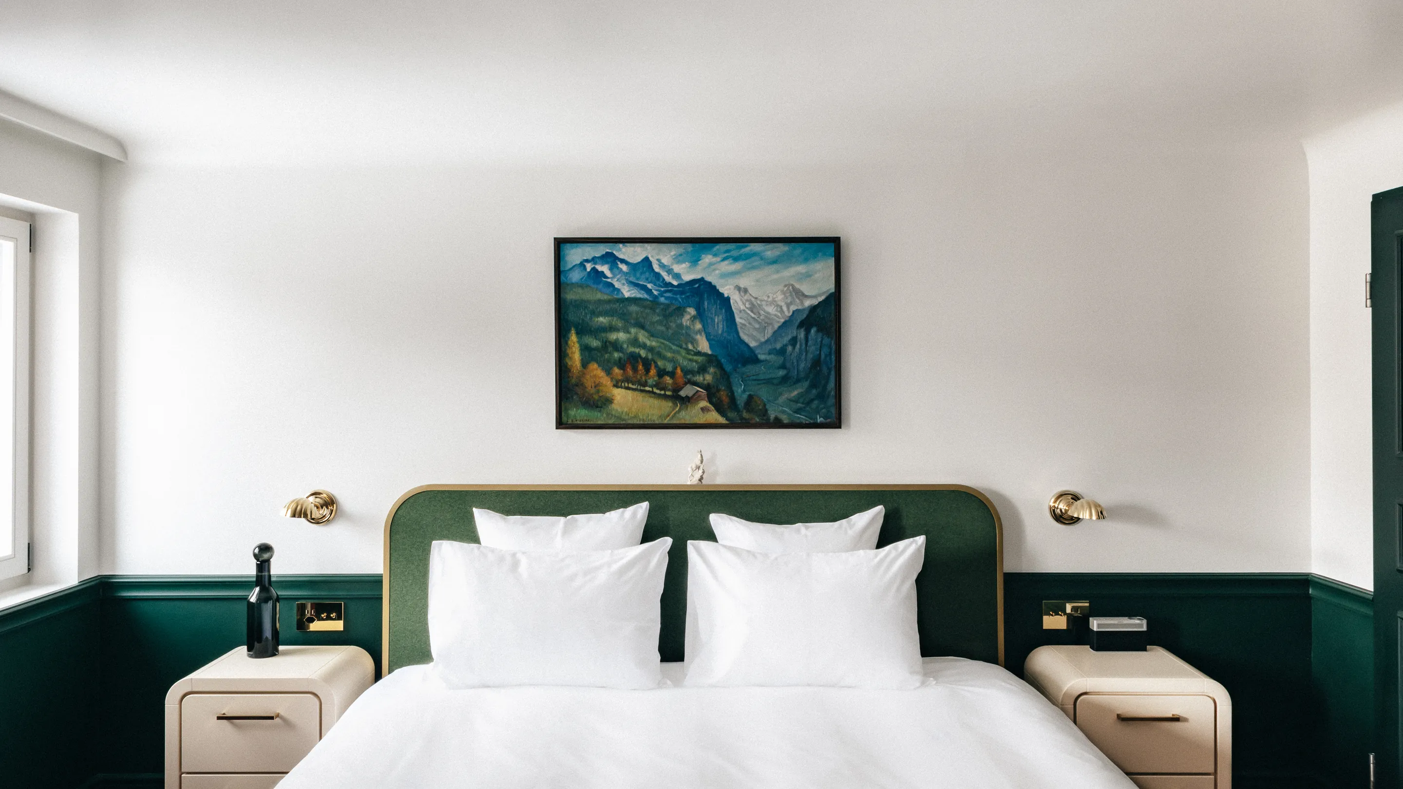 Minimalist bedroom with white bedding, a green upholstered headboard, two beige nightstands, and a framed mountain landscape painting above the bed.