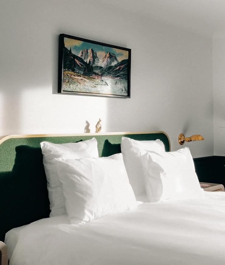 Sunlit bed with white pillows, green headboard, landscape painting, and a wall-mounted wooden reading lamp in a minimalist room.
