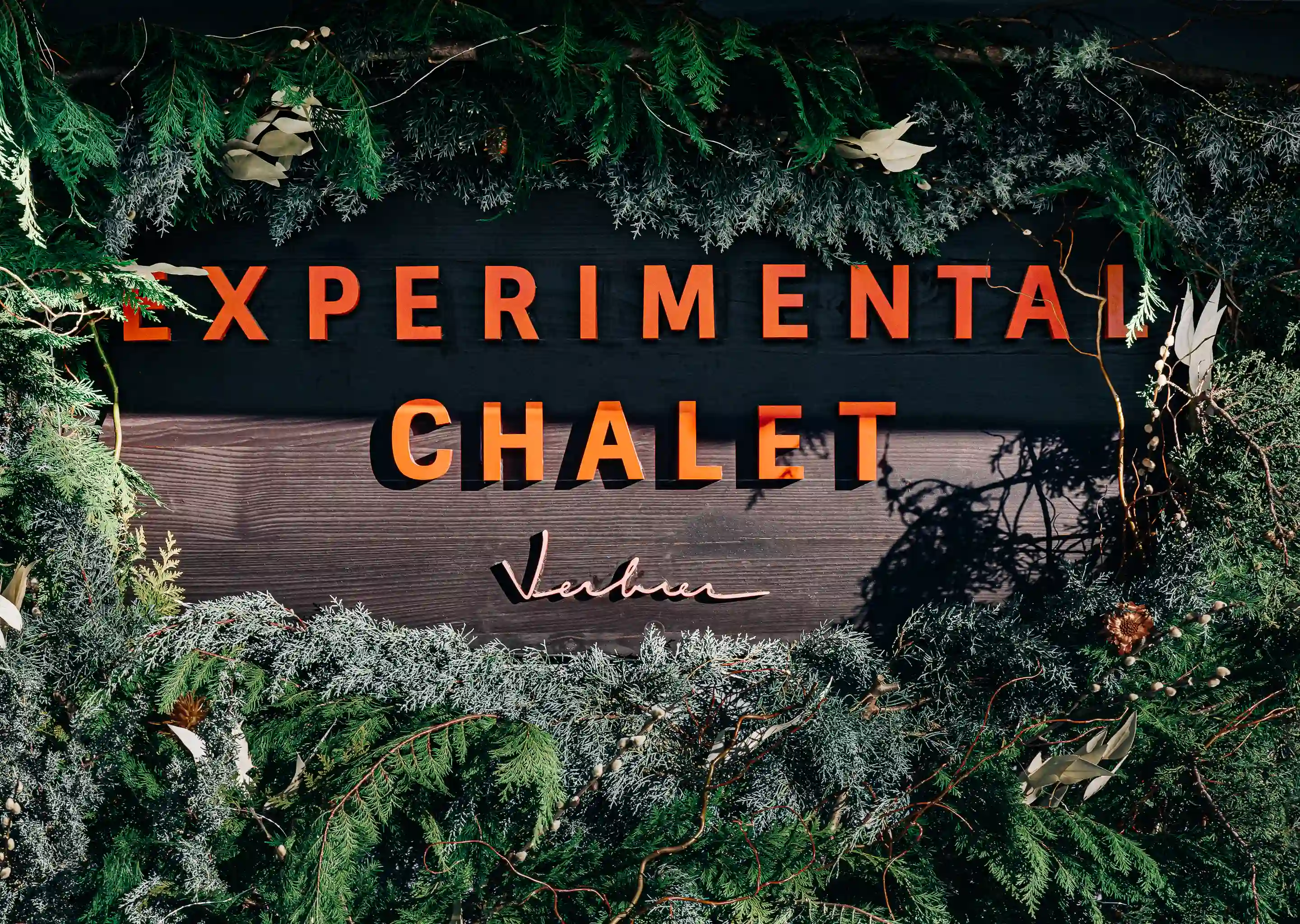 Wooden sign reading 'Experimental Chalet Verbier' surrounded by green and frosted pine branches and foliage.