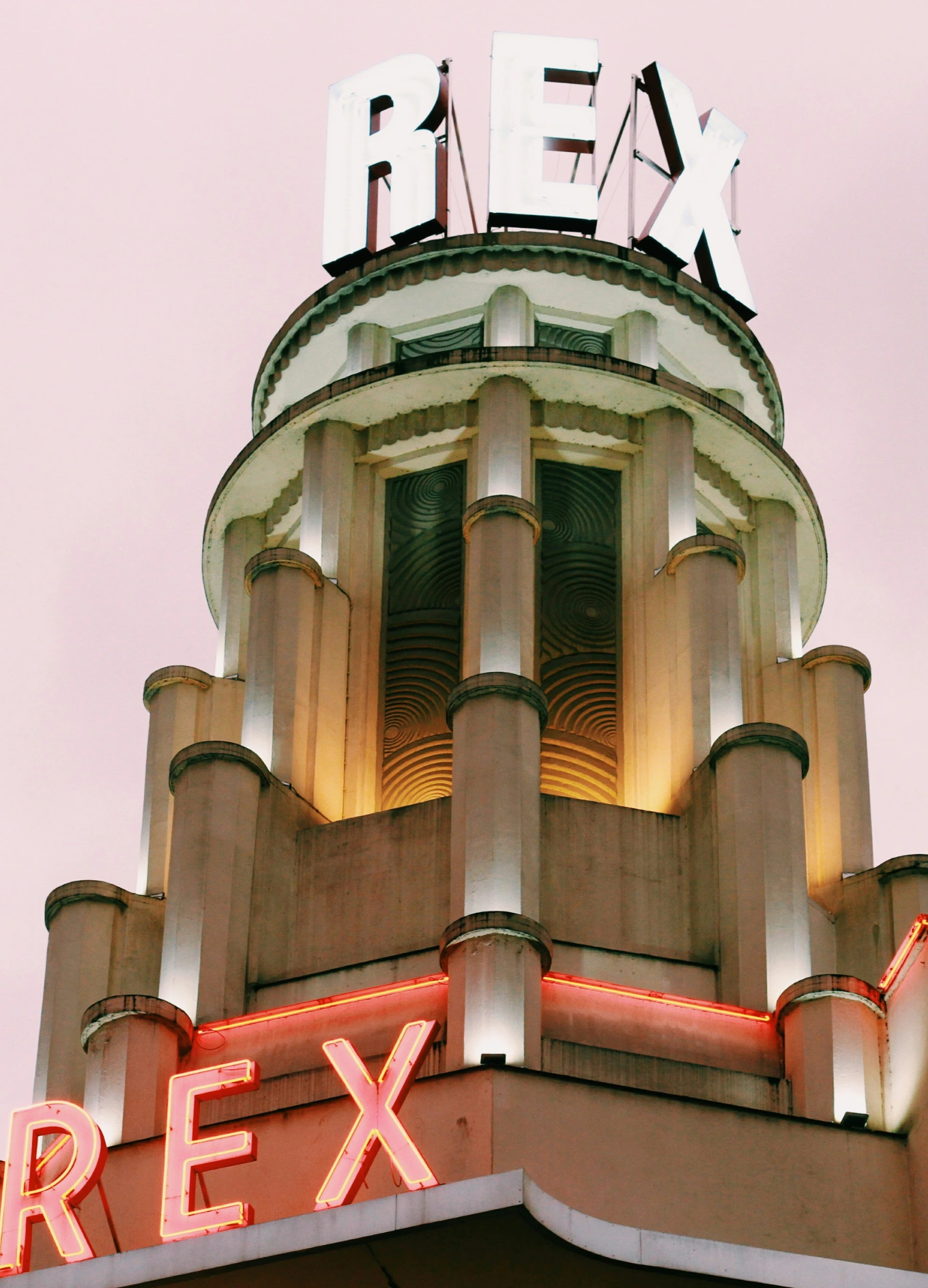 Architectural tower of Hotel Rex lit with neon letters spelling 'REX' against a pink sky.