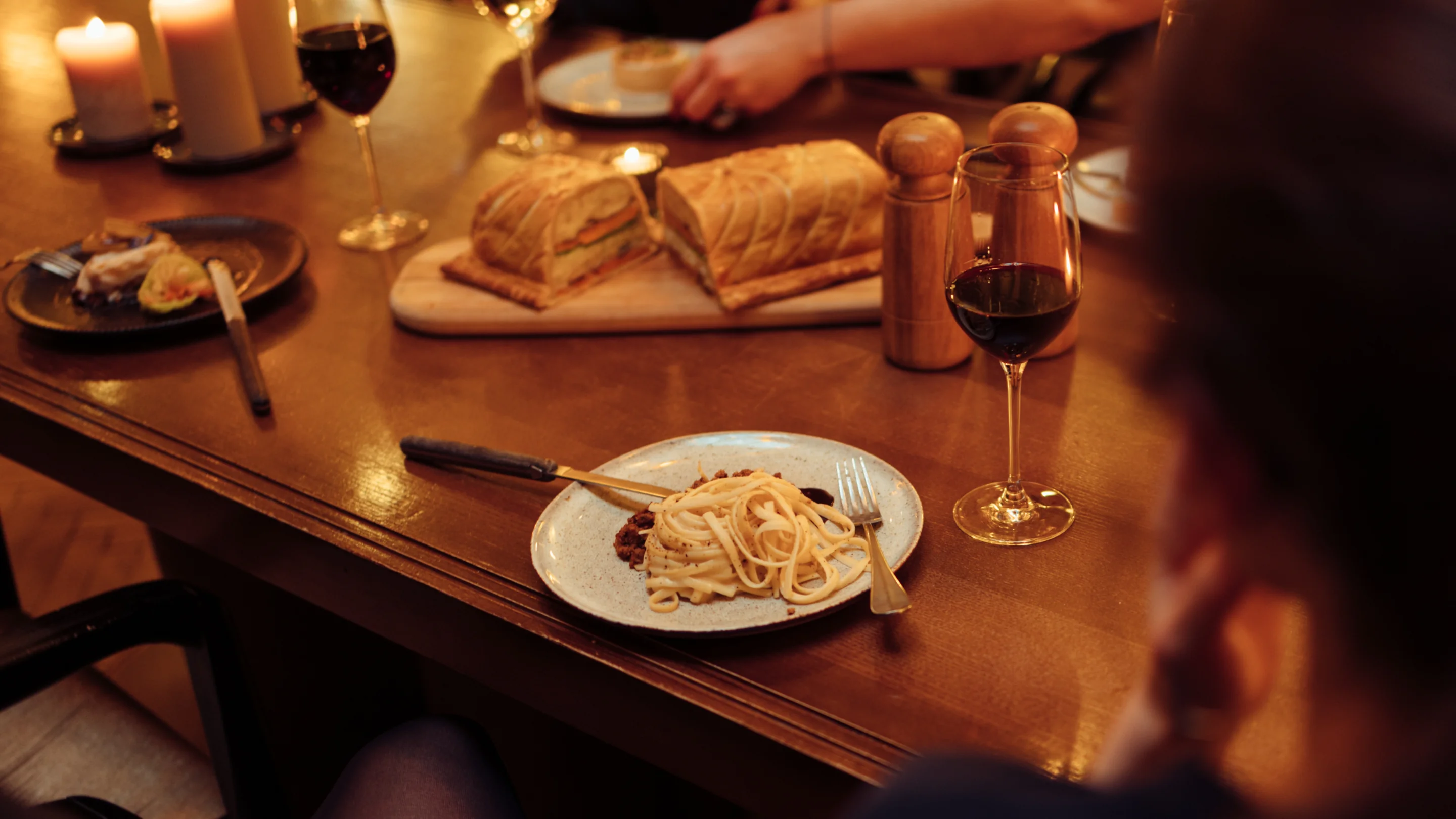 Mesa con platos de pasta, pan, vasos de vino tinto, velas y personas sentadas a su alrededor.