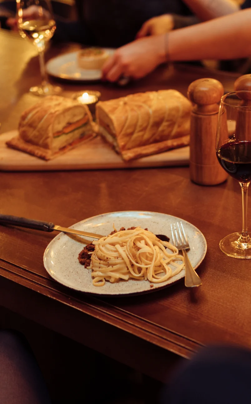 Plato de espaguetis con salsa junto a una copa de vino tinto y un molinillo de pimienta de madera sobre una mesa de madera con una mano que busca comida en el fondo.