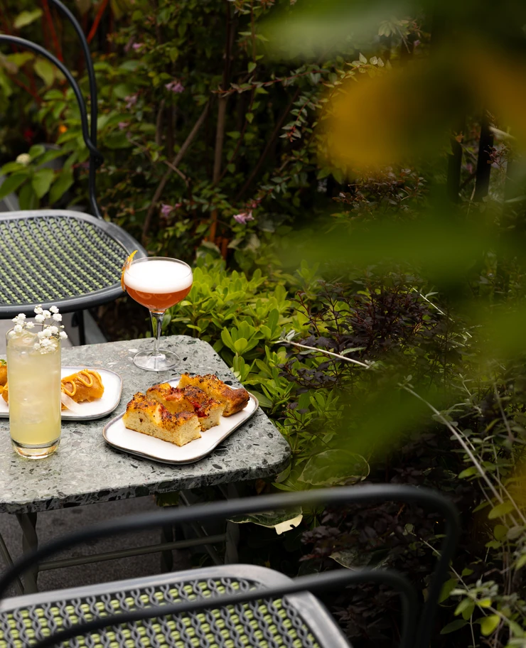 Outdoor table with two cocktails and plates of pastries surrounded by greenery and metal chairs.