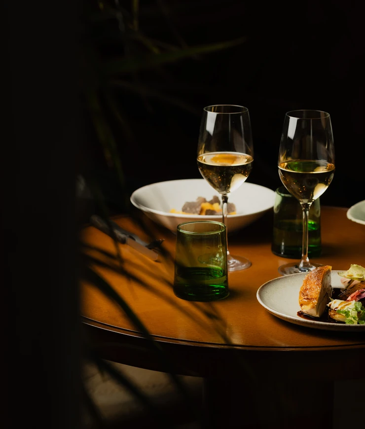 Two glasses of white wine, green water glasses, and plates with gourmet food on a wooden table in a dimly lit setting.