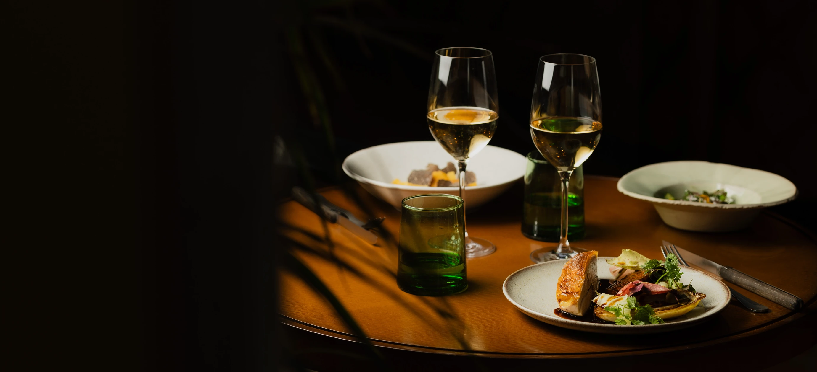 Fine dining table with two glasses of white wine, plates of gourmet food, and green glass tumblers on a wooden surface.
