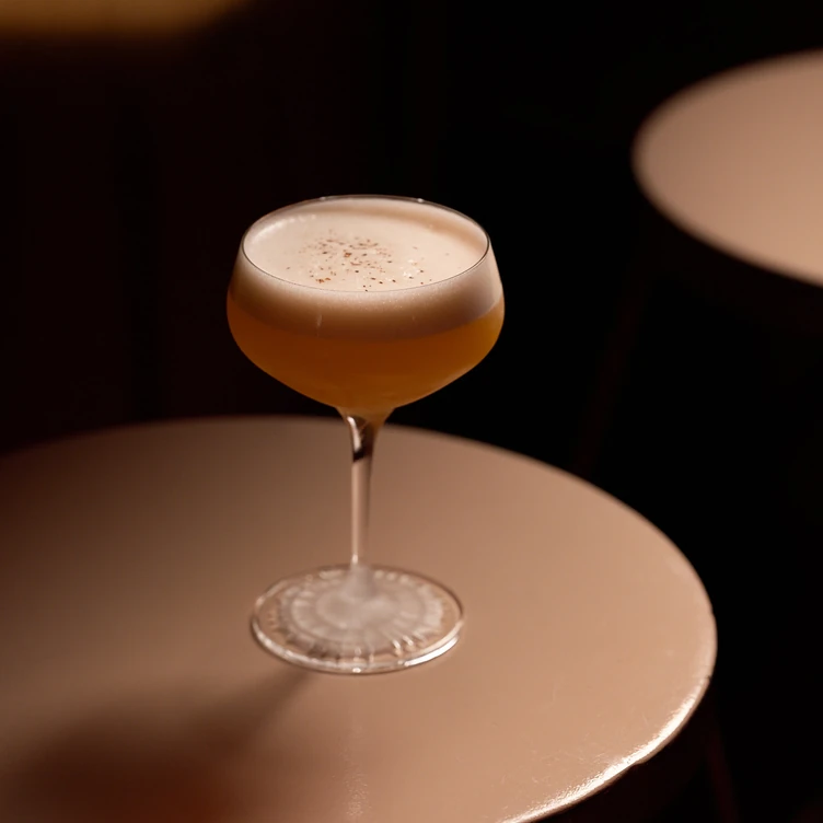 Cocktail with frothy top and sprinkled garnish in a coupe glass on a round beige table in dim lighting.