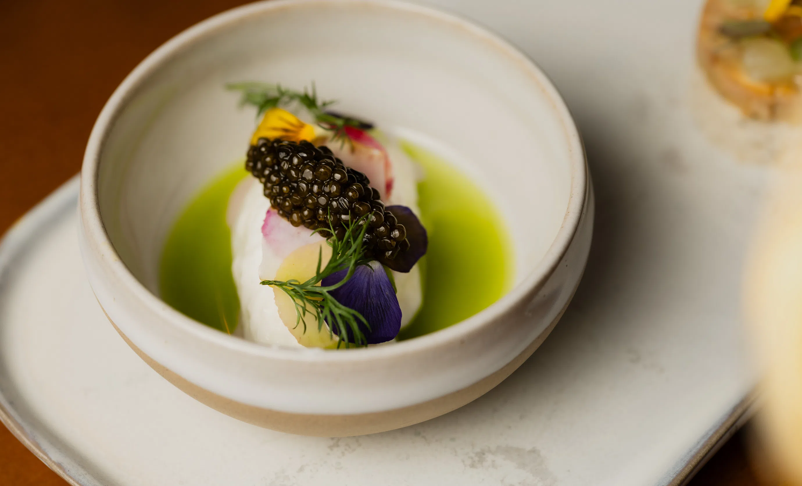 Gourmet dish with black caviar, edible flowers, and herbs on a white mousse base in a green sauce served in a white bowl.
